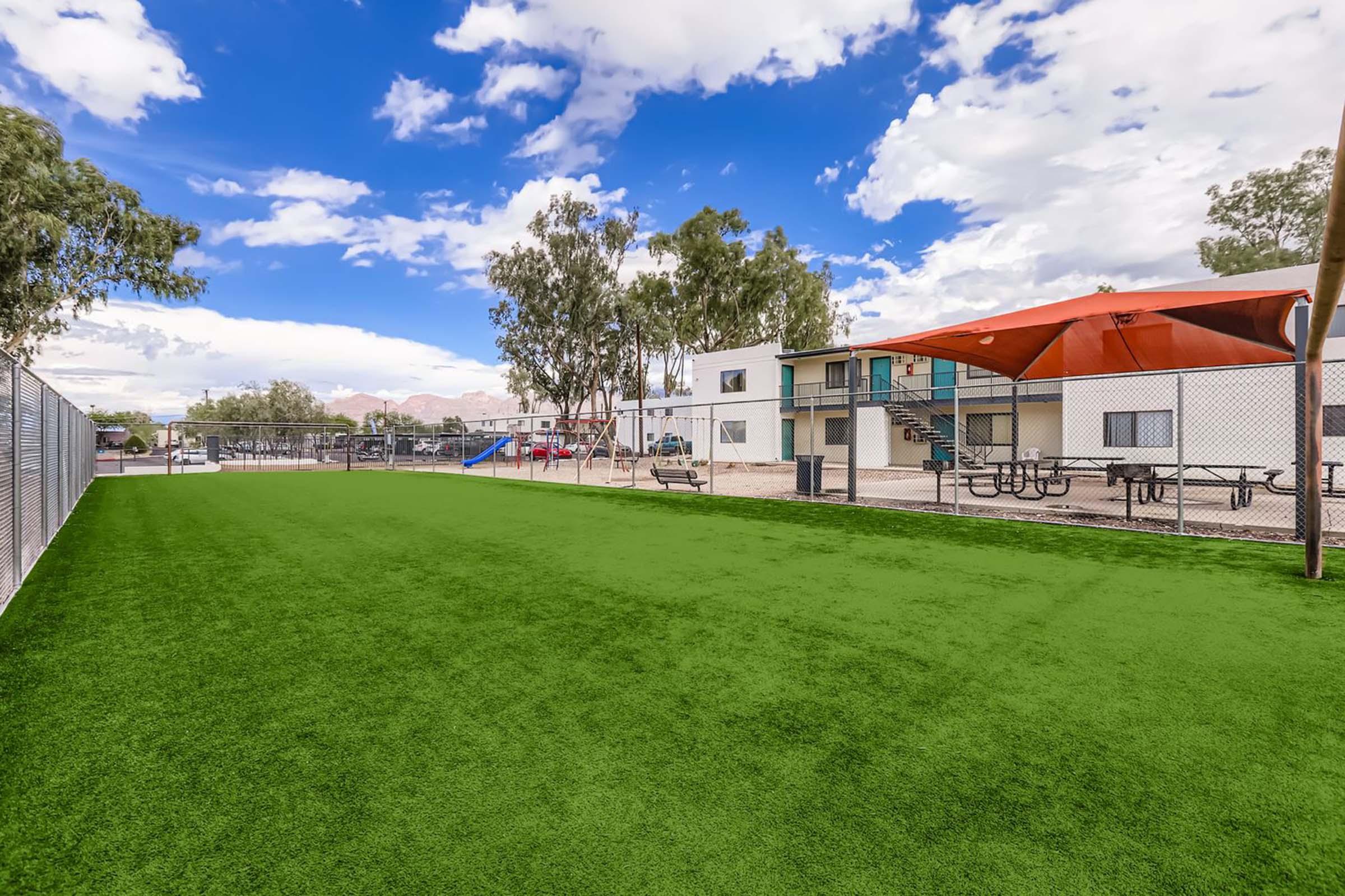 A bright and spacious outdoor area with artificial grass, featuring a picnic area with tables and a playground in the background. The setting includes several trees and a building with balconies, under a partly cloudy blue sky.
