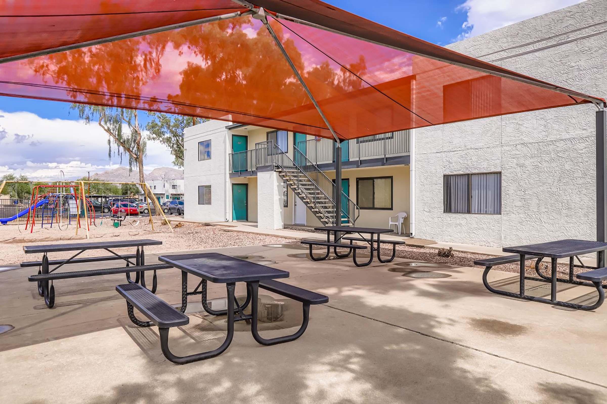 Outdoor area featuring a shaded picnic setup with black tables and benches, surrounded by a playground and a multi-story building with balconies. The space is well-lit, with a vibrant orange canopy providing shade amidst a clear sky.