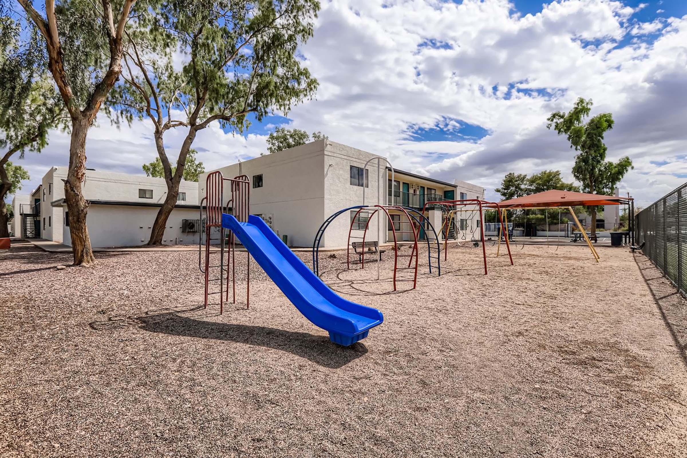 A playground area featuring a blue slide, red swing set, and climbing frame. The ground is covered in gravel, surrounded by trees and a building in the background under a partly cloudy sky.