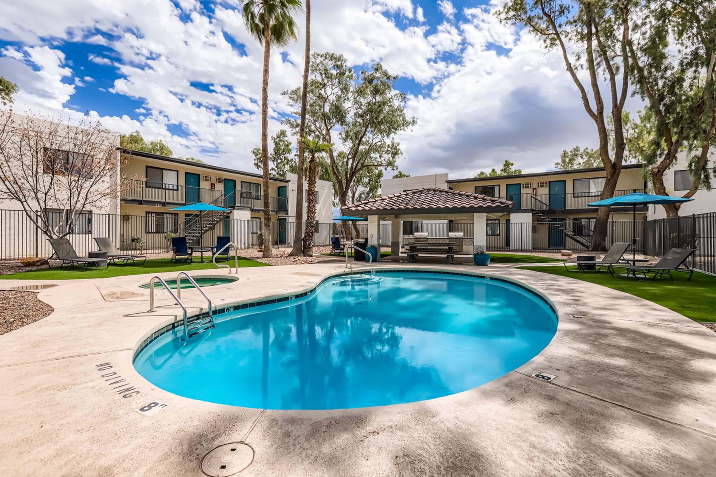 A clear, inviting swimming pool shaped like an oval, surrounded by lounge chairs and umbrellas. In the background, several modern apartment buildings with palm trees and a blue sky dotted with clouds enhance the relaxing atmosphere.