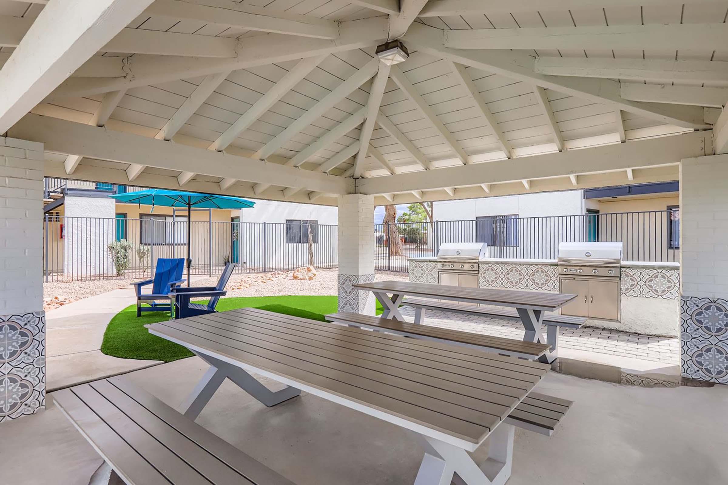 A spacious outdoor pavilion with a peaked ceiling, featuring picnic-style tables and stainless steel grills. Sunlight streams in through the open sides, and a small grassy area is visible. A blue lounge chair sits nearby under an umbrella, creating an inviting atmosphere for gatherings and barbecues.