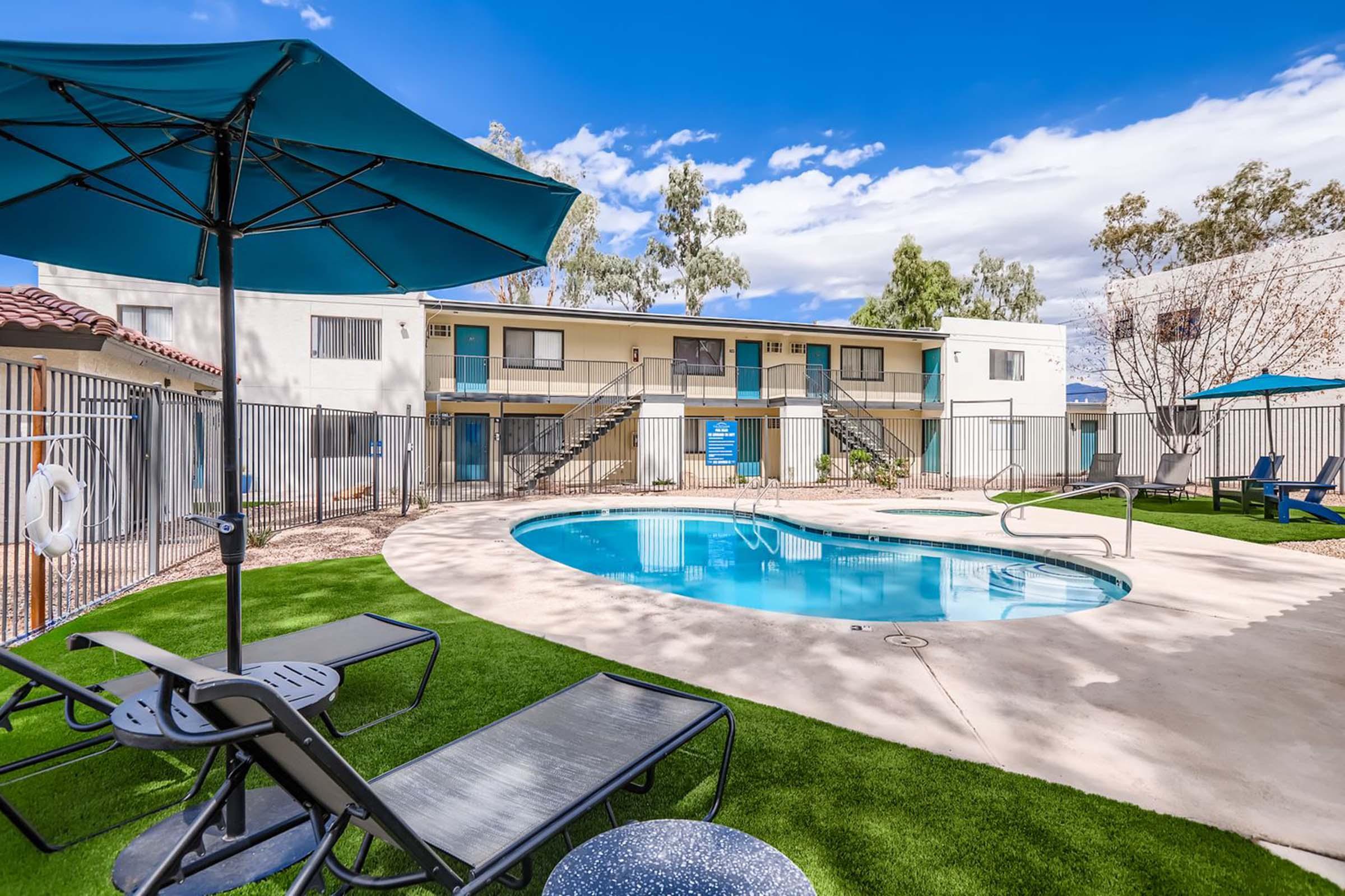 A clear blue swimming pool surrounded by lounge chairs and umbrellas, with two multi-story apartment buildings in the background. Lush green grass borders the pool area, set against a sunny sky with a few clouds. The scene conveys a relaxing outdoor space ideal for leisure and recreation.