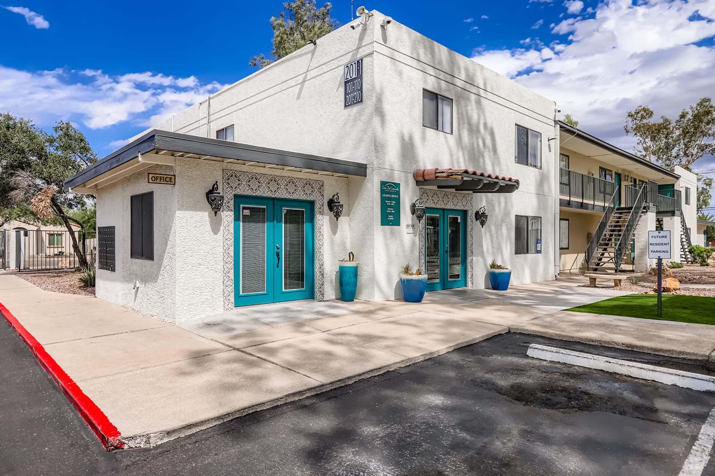 A white two-story building with turquoise doors and windows. The front features decorative elements and planters. A sign indicates an office. The driveway is paved, and there is a parking area in front. The background includes trees and a blue sky with clouds.