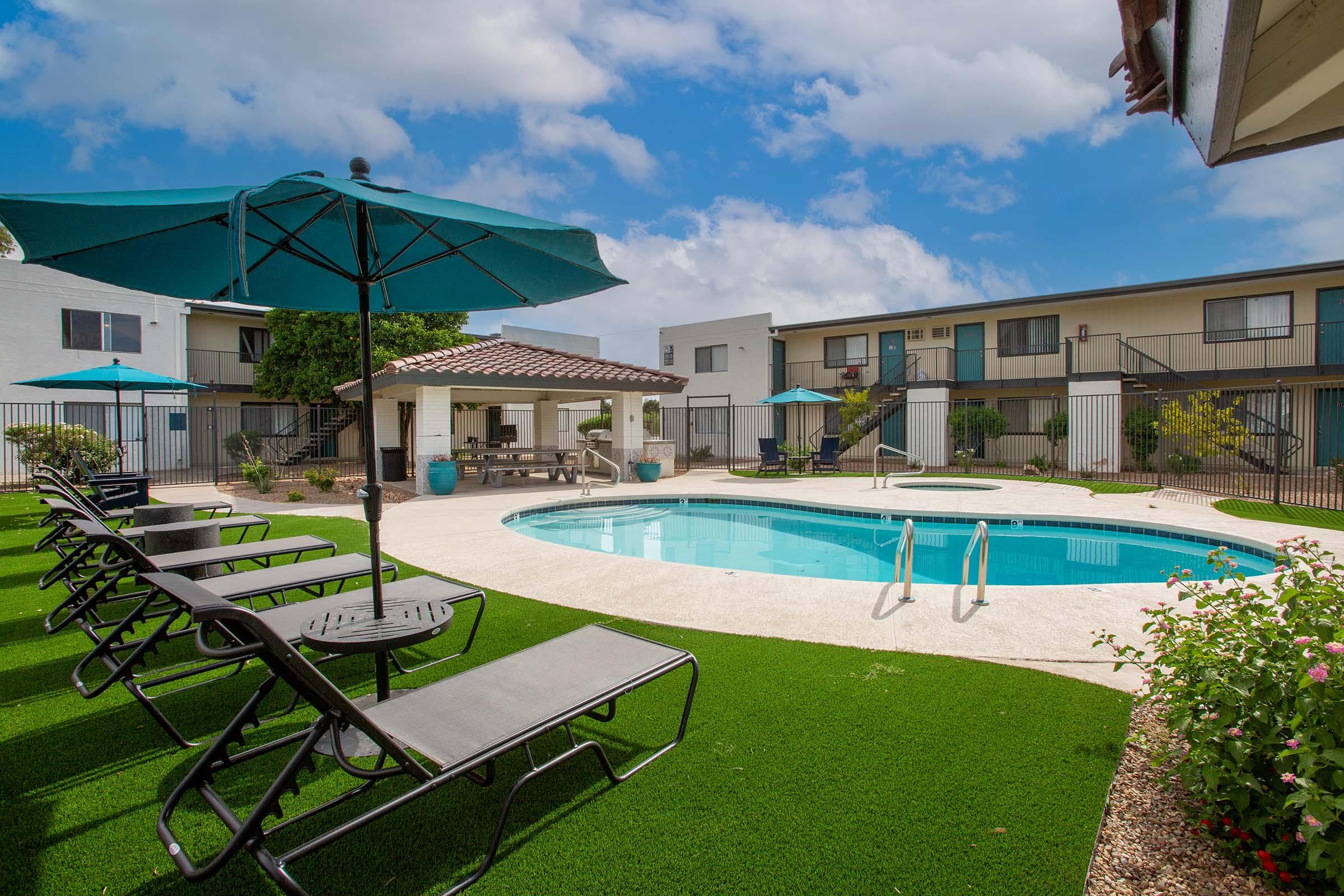 A sunny pool area surrounded by lounge chairs and umbrellas, featuring a clear blue pool and landscaped green grass. In the background, white buildings with balconies create a relaxing atmosphere, and a shaded gazebo offers a space for gatherings. The sky is partly cloudy, enhancing the inviting ambiance.