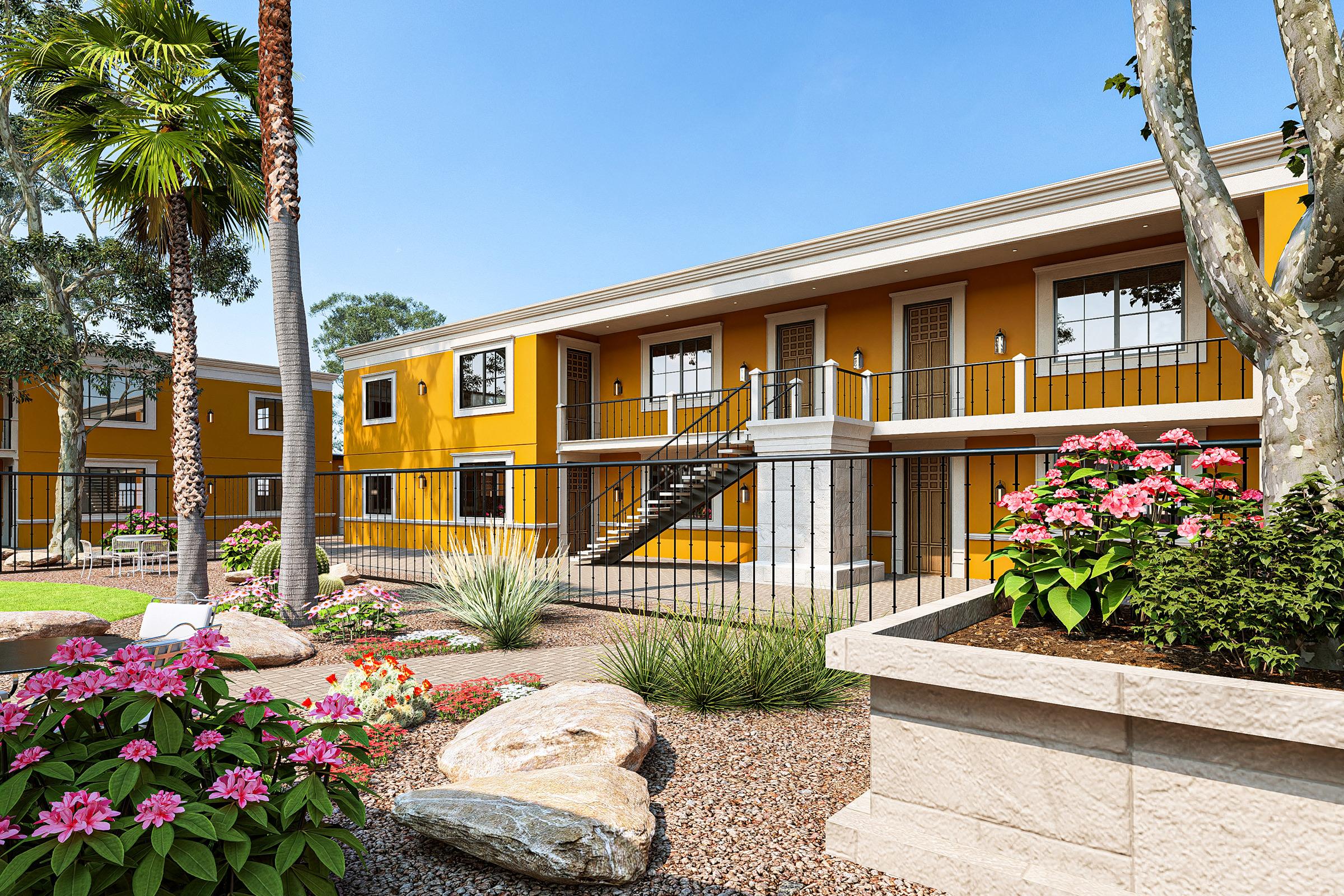 A well-maintained apartment complex featuring bright yellow buildings, surrounded by lush landscaping with colorful flowers and palm trees. The outdoor area includes decorative stones and plants, with a central pathway leading to the staircases of the buildings under a clear blue sky.