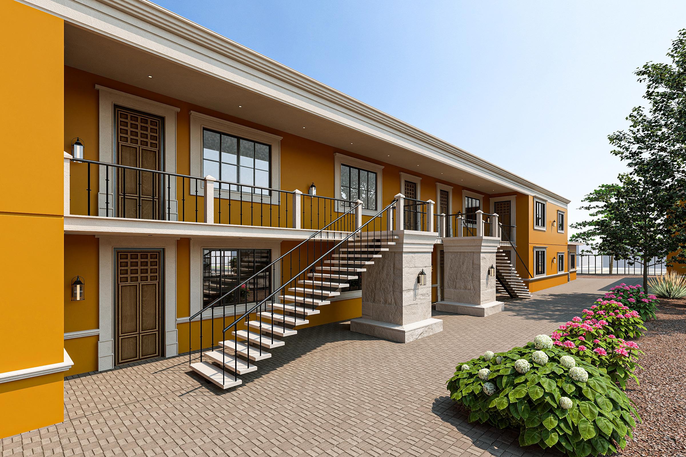 A view of a modern, two-story building with yellow exterior walls, featuring balconies with railings and staircase access. A well-maintained garden with flowering plants is in the foreground, and there are trees lining the side of the property under a clear blue sky.