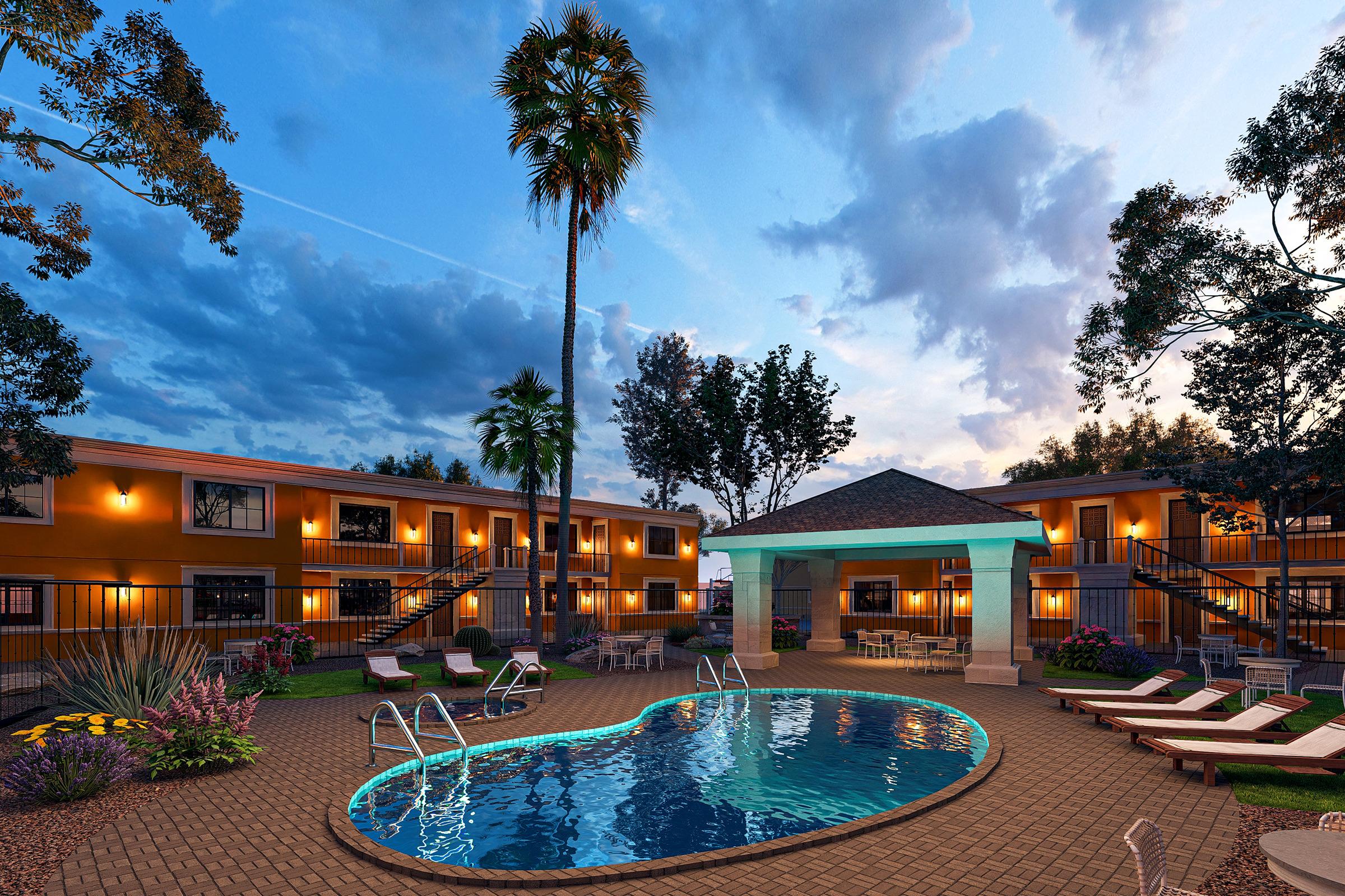 A scenic view of a hotel courtyard featuring a shimmering pool surrounded by lounge chairs and palm trees. The two-story buildings are painted in warm colors, with a gazebo providing shade. The sky is dusk, displaying a mix of blue and purple hues with scattered clouds. Lush, green landscaping enhances the tranquil atmosphere.