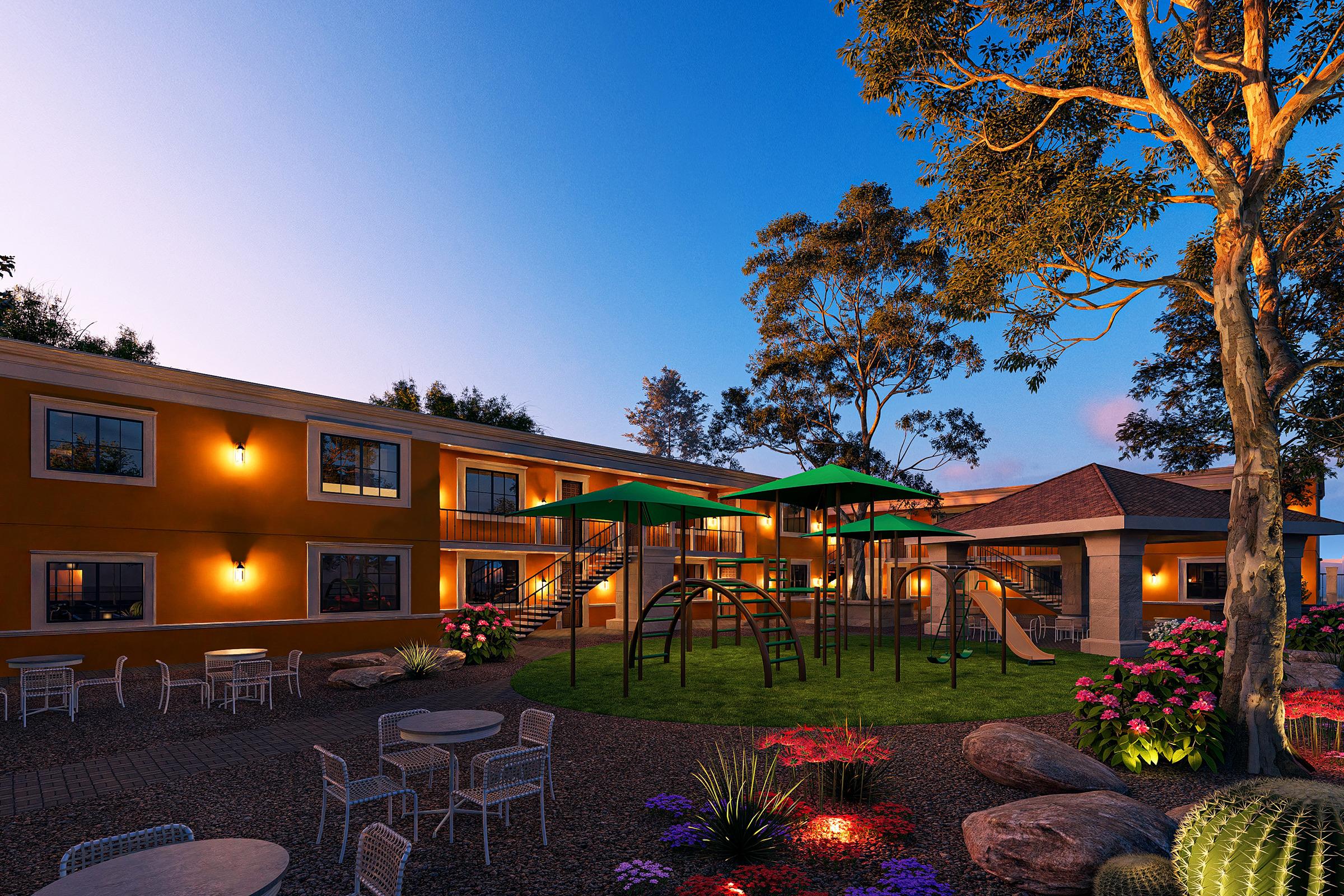 A colorful outdoor area featuring a playground with green canopies, surrounded by flower beds and rocky landscaping. In the background, a two-story building with bright orange walls and large windows is illuminated by warm lights, under a clear blue sky during dusk.