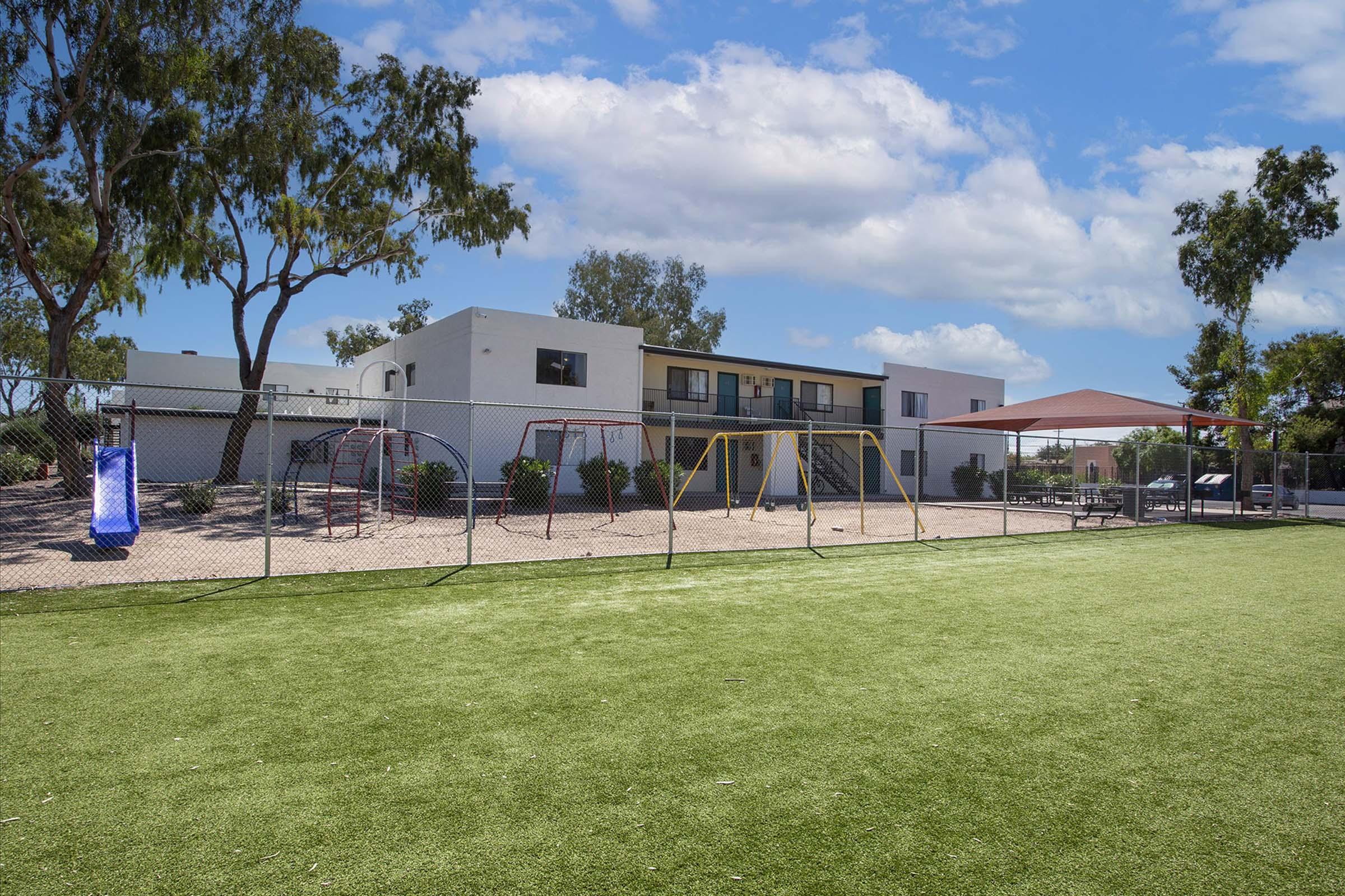 A playground with swings and slides on a grassy field, surrounded by a chain-link fence. In the background, there is a low-rise building with several windows and trees providing shade. The sky is partly cloudy, creating a bright and inviting atmosphere.