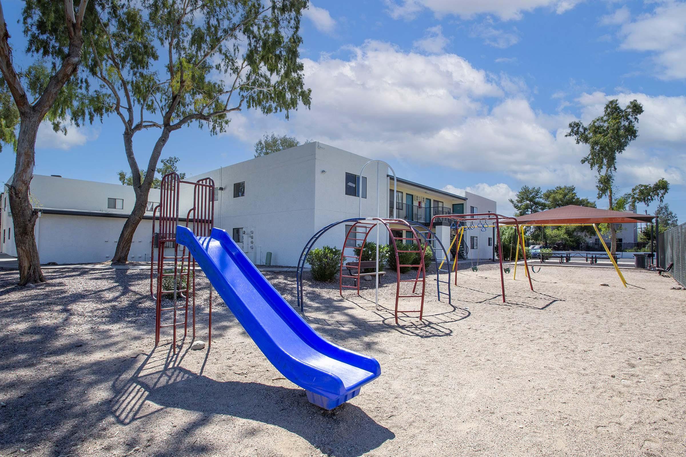 A playground featuring a blue slide, swings, and a shaded structure surrounded by gravel and trees. In the background, there are white buildings under a partly cloudy sky.