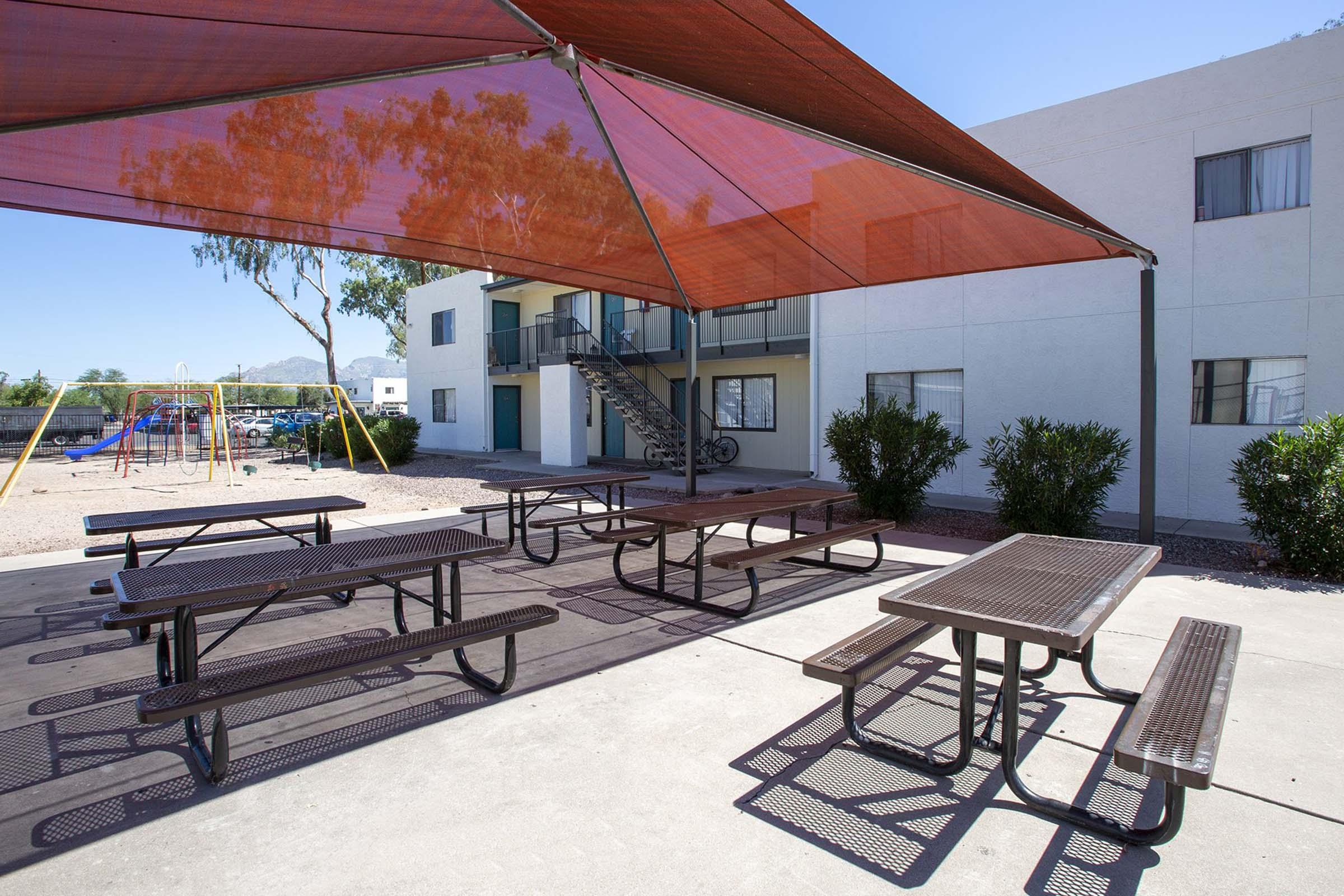 A shaded outdoor area with multiple metal picnic tables, surrounded by greenery. In the background, an apartment building with balconies and a staircase is visible. A playground structure with swings can be seen to the side. The sky is clear and blue, suggesting a sunny day.