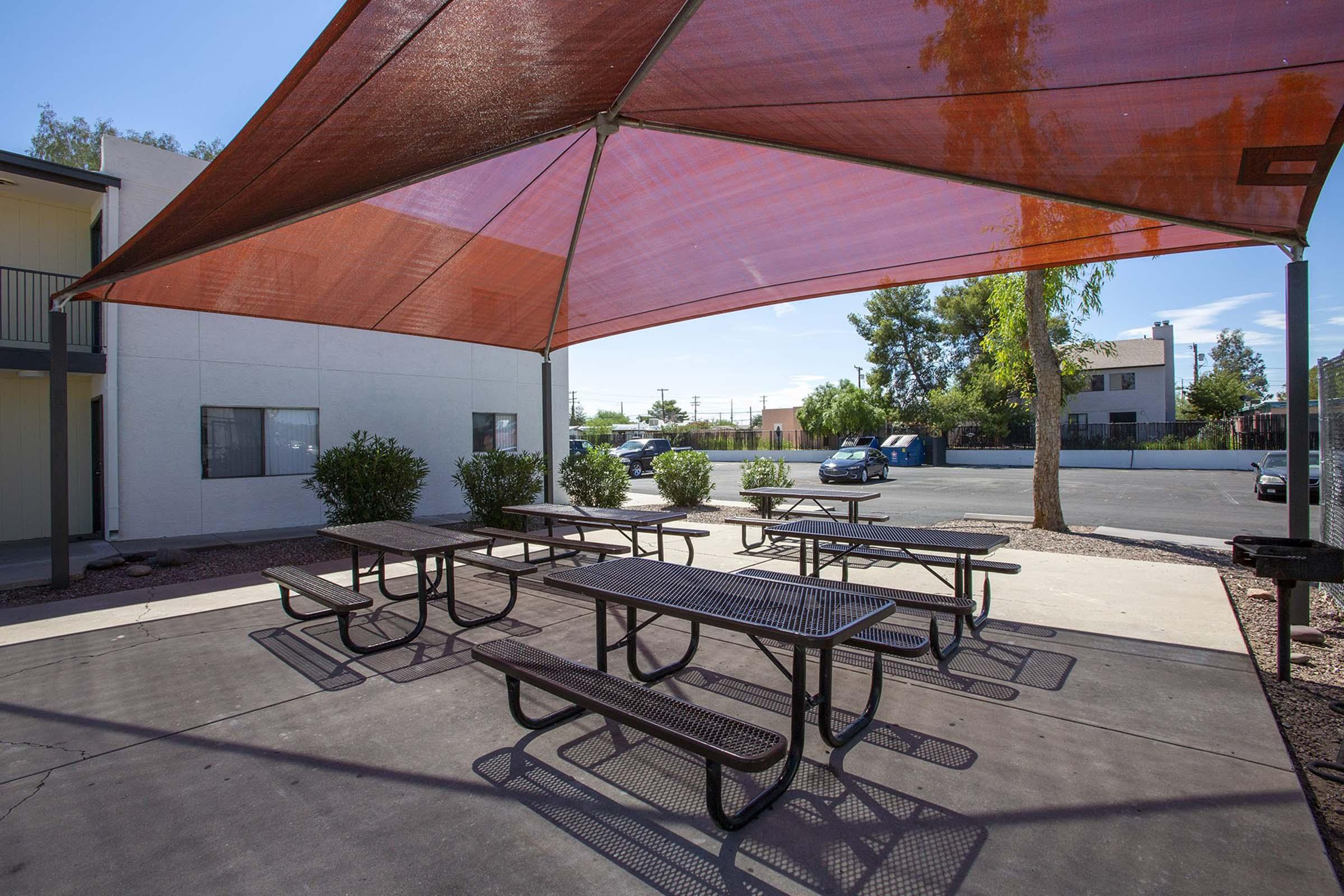 Outdoor area with multiple black picnic tables shaded by a large, orange canopy. Surrounding the tables are landscaped bushes, and in the background, there are parked cars and buildings. The scene is well-lit and appears to be a communal space for gatherings or meals.