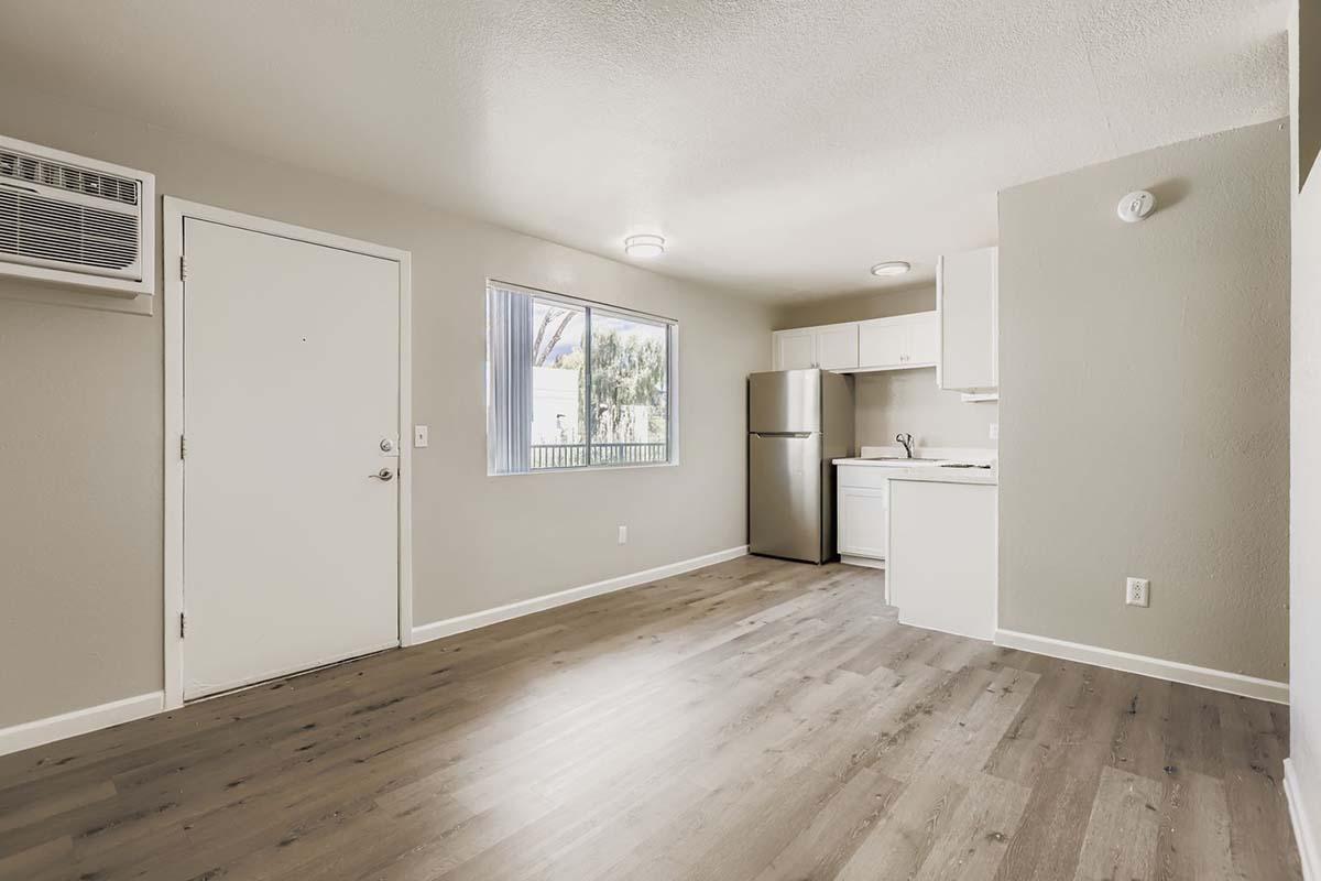 Interior of a small, modern apartment with light gray walls and wooden flooring. The space includes a kitchen area with a white refrigerator, a sink, and a window letting in natural light. A door leads outside, and an air conditioning unit is mounted on the wall. The overall ambiance is bright and airy.