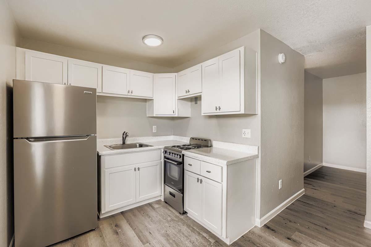 Modern kitchen featuring white cabinetry, a stainless steel refrigerator, and a matching stove and oven. The countertops are light-colored, and there's a sink with a faucet. The walls are painted in a neutral tone, and the flooring is a wood-like material. The layout is compact and functional.