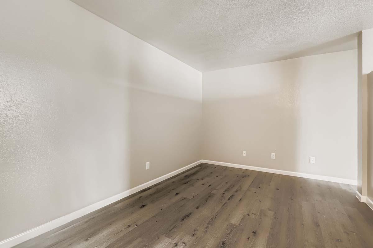 Empty room with light gray painted walls and dark wooden flooring. The space features no furniture and has white electrical outlets on the walls. Natural light illuminates the room, creating a bright and spacious appearance.