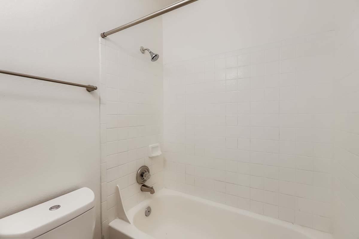 A clean, white bathroom featuring a shower area with tiled walls, a bathtub with a rim, and a toilet. The showerhead is mounted on the wall, and there are towel bars on either side. The overall design is modern and minimalist with a bright, neutral color scheme.