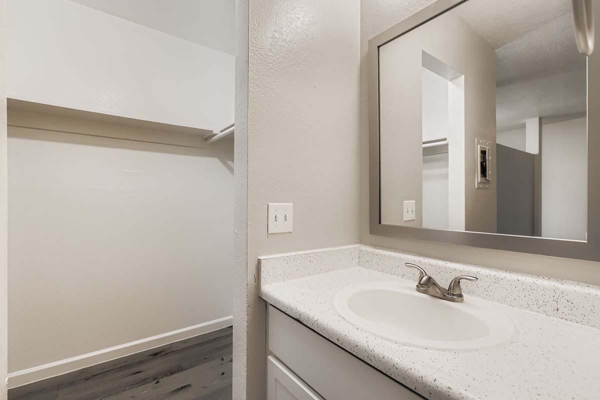 A modern bathroom featuring a clean white countertop with a single sink and a rectangular mirror above it. To the left, there is an open closet space with shelving, and the walls are painted in light neutral tones, creating a bright and airy atmosphere. The flooring appears to be laminate in a dark shade.