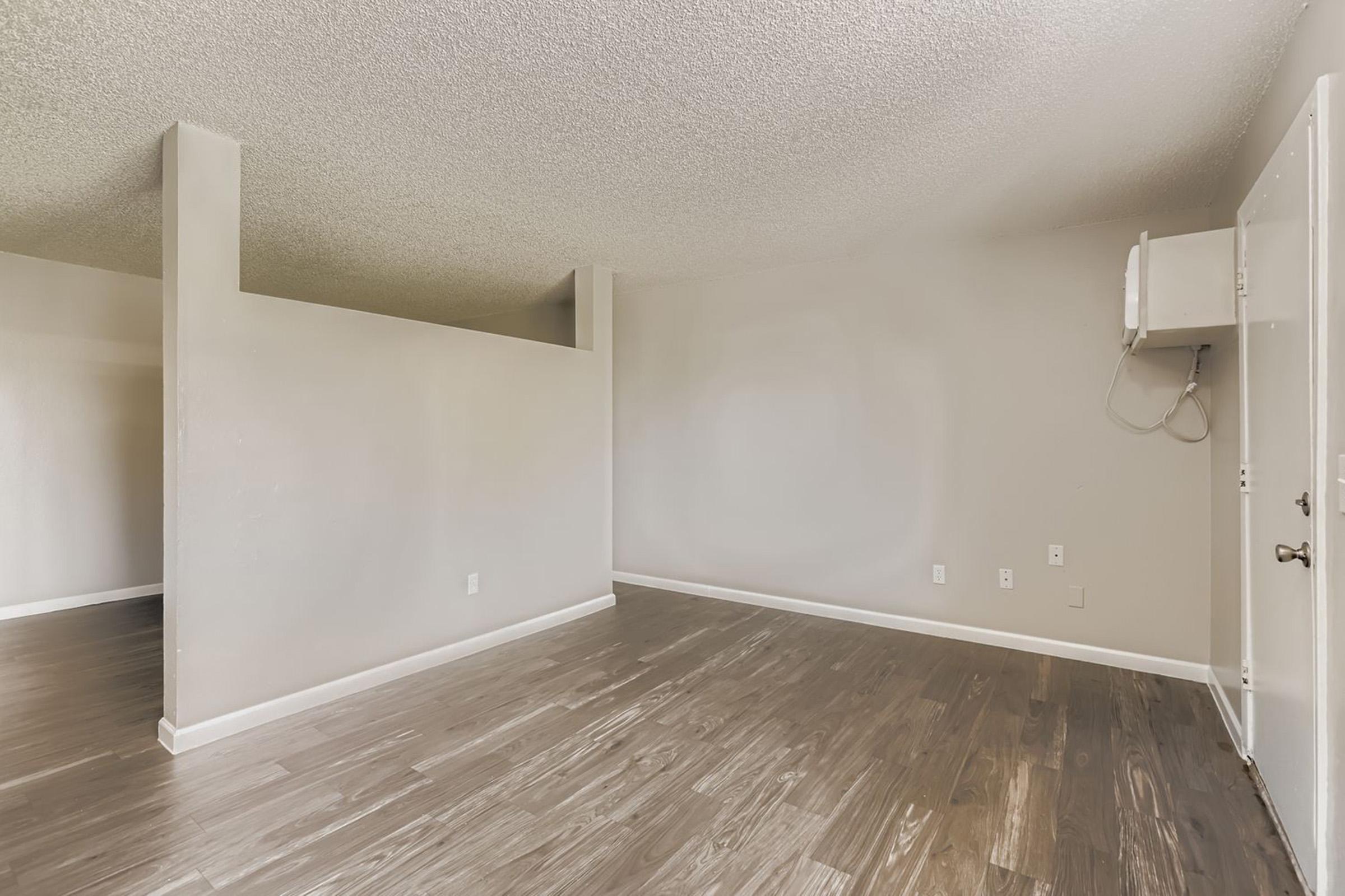 An empty room with light-colored walls and a textured ceiling. The flooring is dark wood, and there is a partial wall separating the space. To the right, there is a door and a small area with wall-mounted equipment. The room is well-lit and has a clean, spacious appearance.