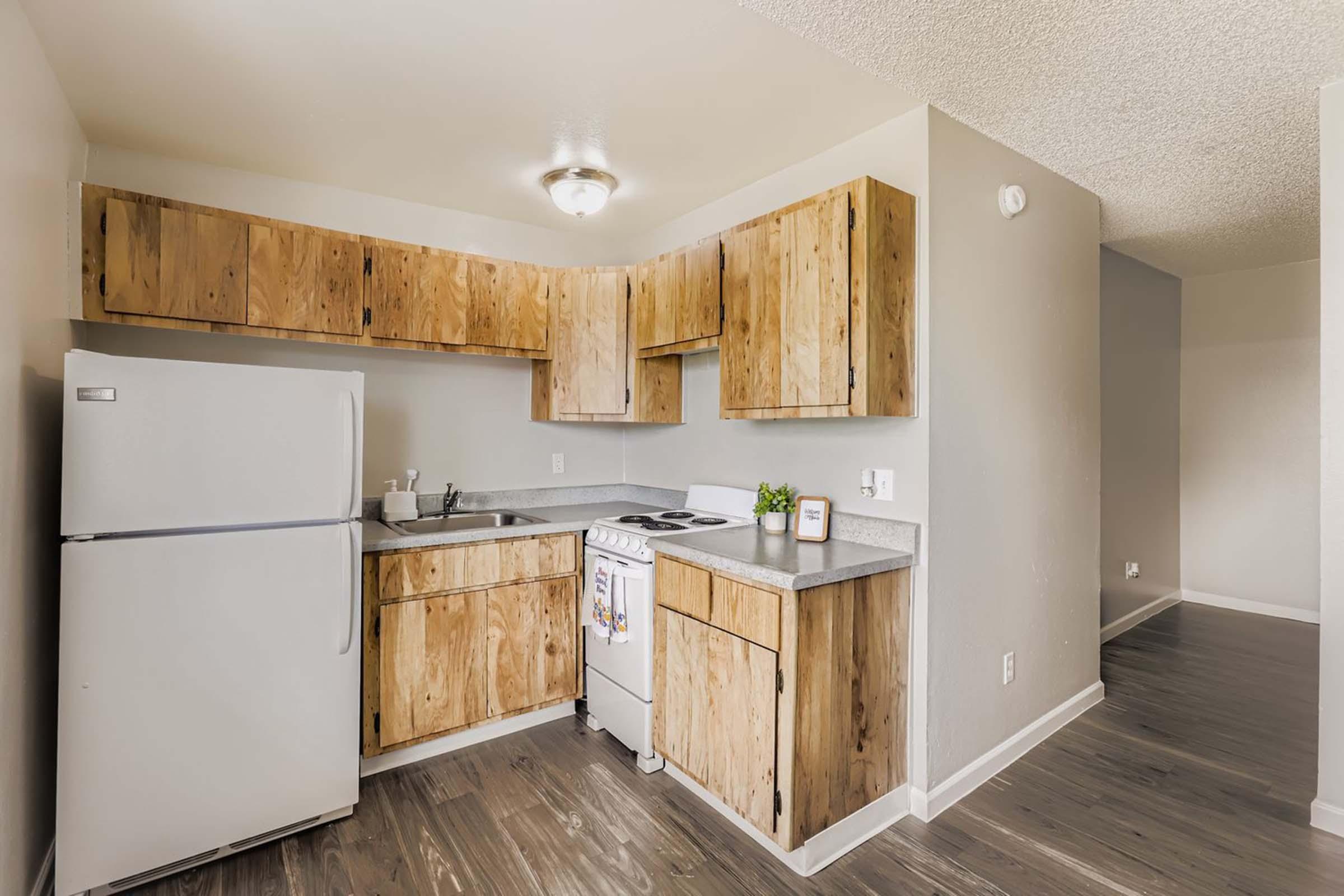 A compact kitchen featuring wooden cabinets, a white refrigerator, a sink, and a stove. The countertops are light gray, and the walls are painted in a neutral tone. The kitchen has modern lighting and a clean, minimalist design. A doorway leads to another room in the background.