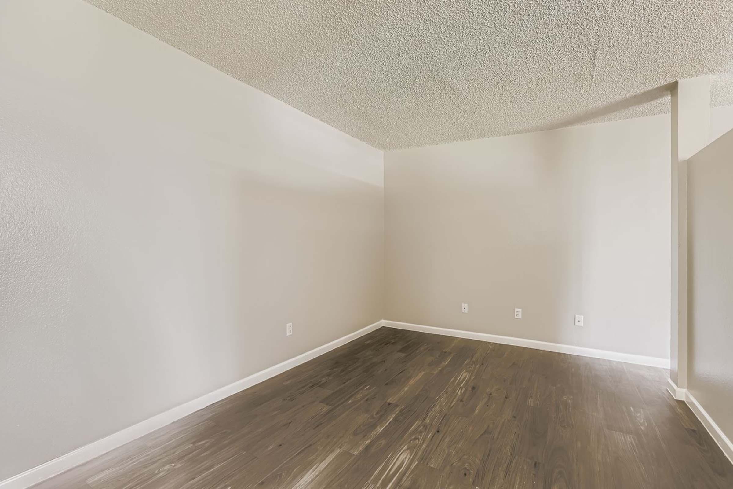 Empty room with light gray walls and a textured ceiling. The floor is covered with dark wood planks. There are no furniture items, providing a spacious and clean aesthetic. The image captures one corner of the room, highlighting the simplicity and openness of the space.