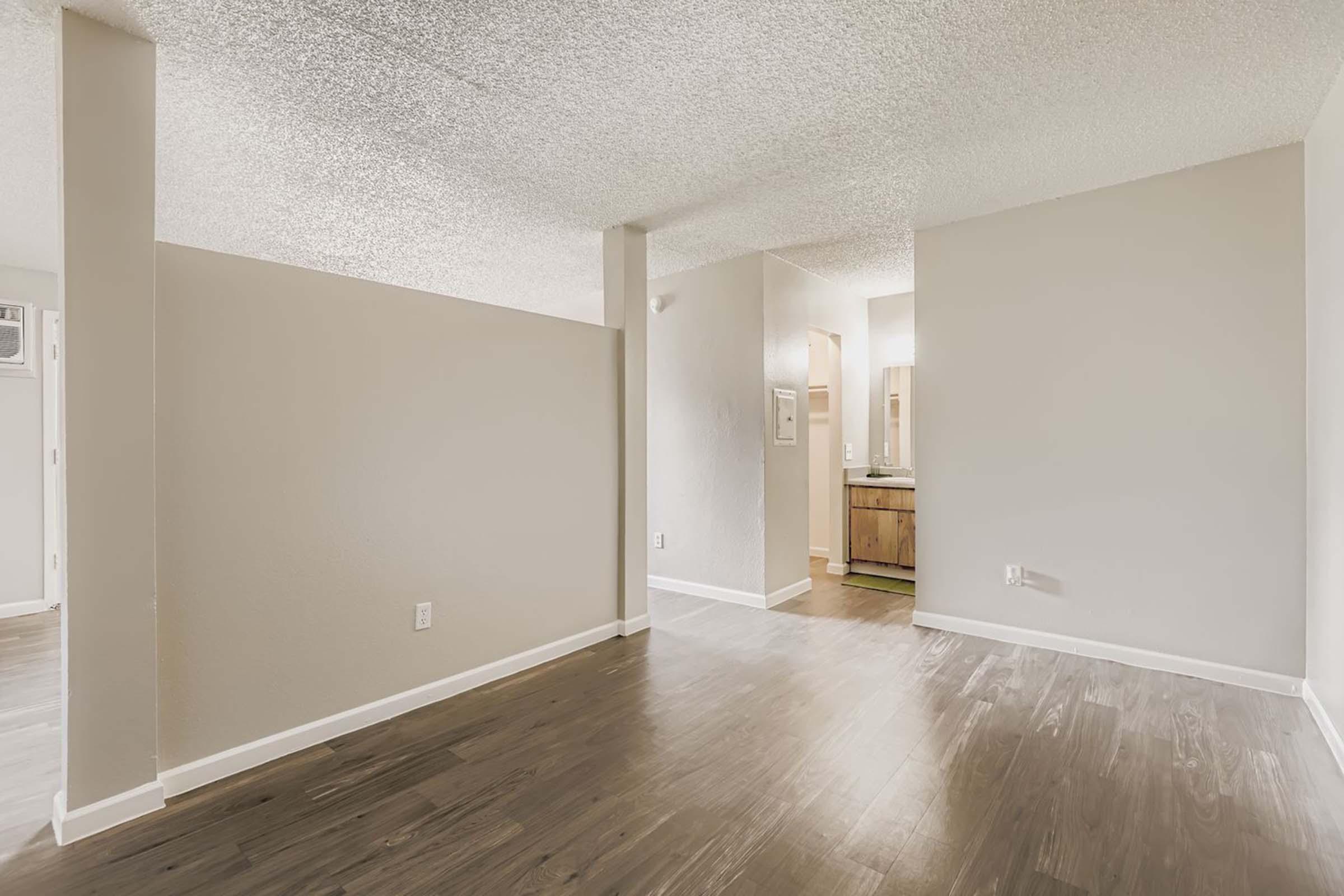 An interior view of a spacious room with light-colored walls and wooden flooring. There's a partition wall to the left, leading to a bright area that may include a bathroom. The overall atmosphere is clean and modern, with ample natural light.