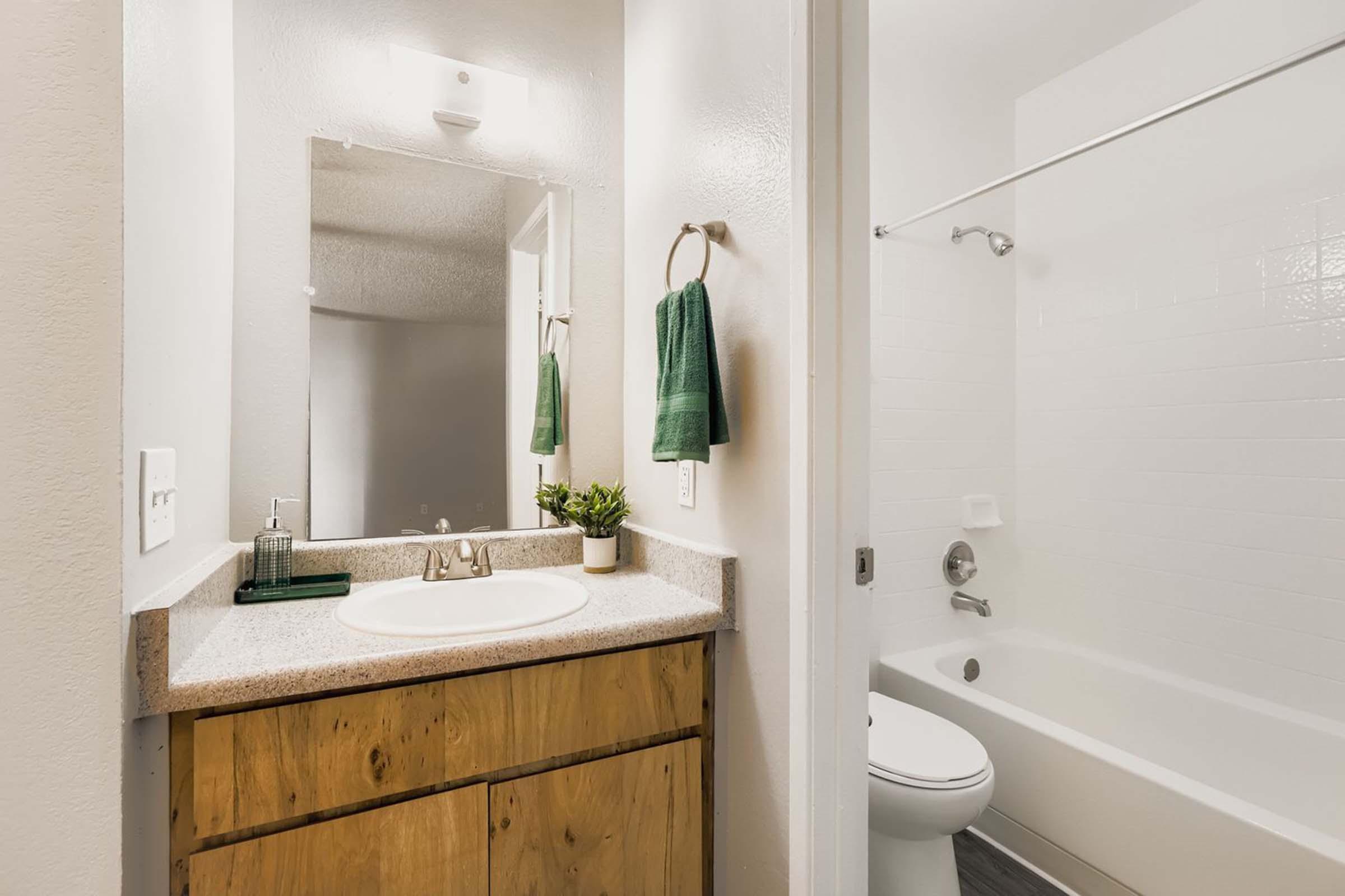 A small bathroom featuring a sink with a mirror above, wooden cabinetry, and a white bathtub with a shower curtain. Green towels hang on a towel rack, and a small potted plant adds a touch of decor. The walls are painted in neutral tones, creating a light and airy atmosphere.