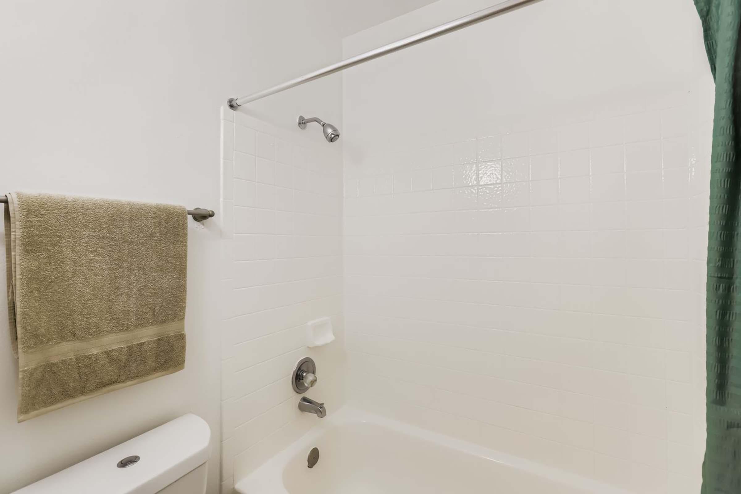 A clean bathroom featuring a shower area with a white tiled wall, a showerhead, and a bathtub. A light green towel hangs on a bar, and a white toilet is visible adjacent to the shower. The overall atmosphere is simple and minimalistic.
