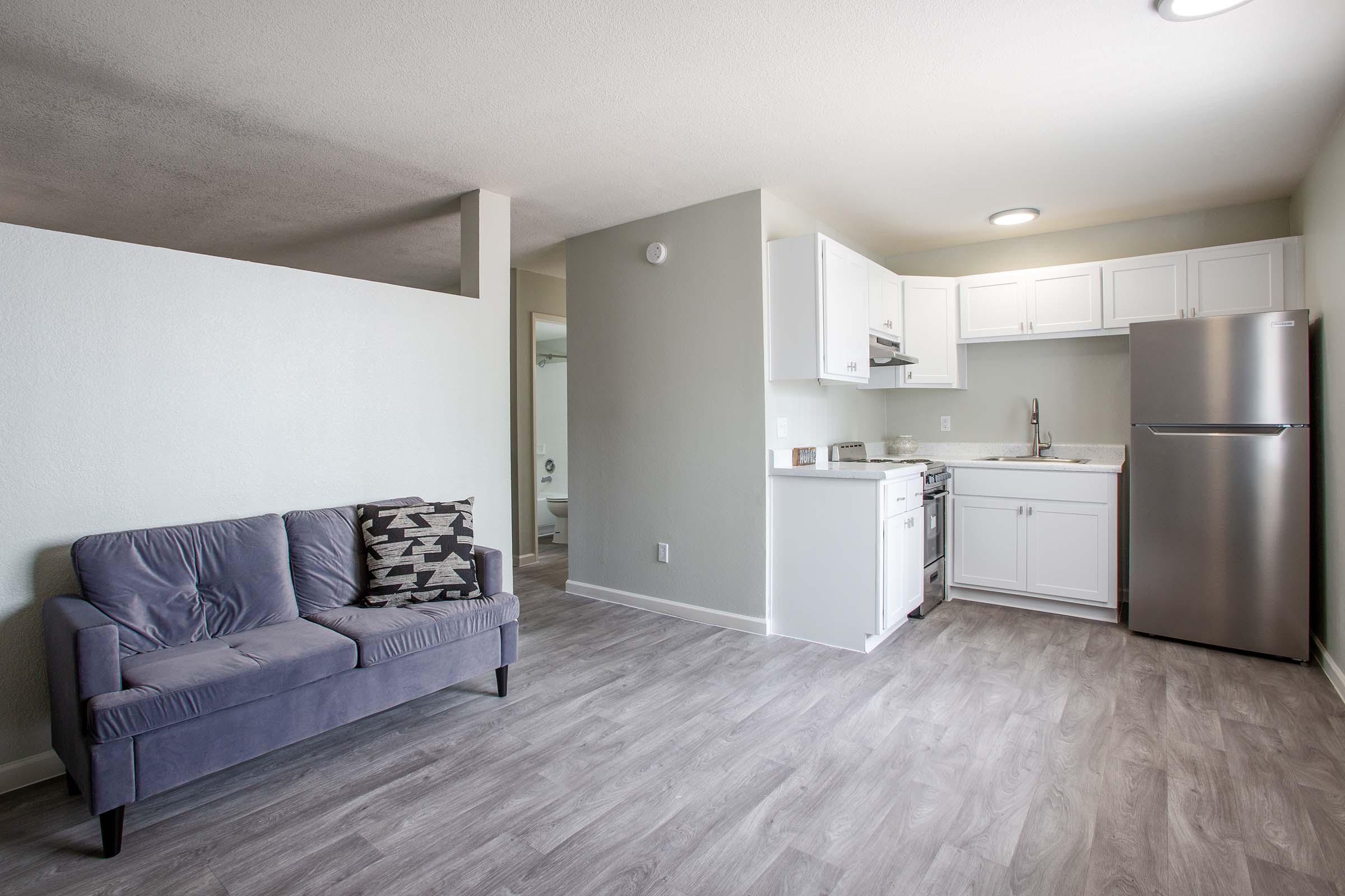 A bright and modern living space featuring a gray sofa with patterned cushions, a small kitchen area with white cabinets, a stainless steel refrigerator, and a stove. The flooring is light gray, and the walls are painted a soft neutral color, creating a welcoming atmosphere.
