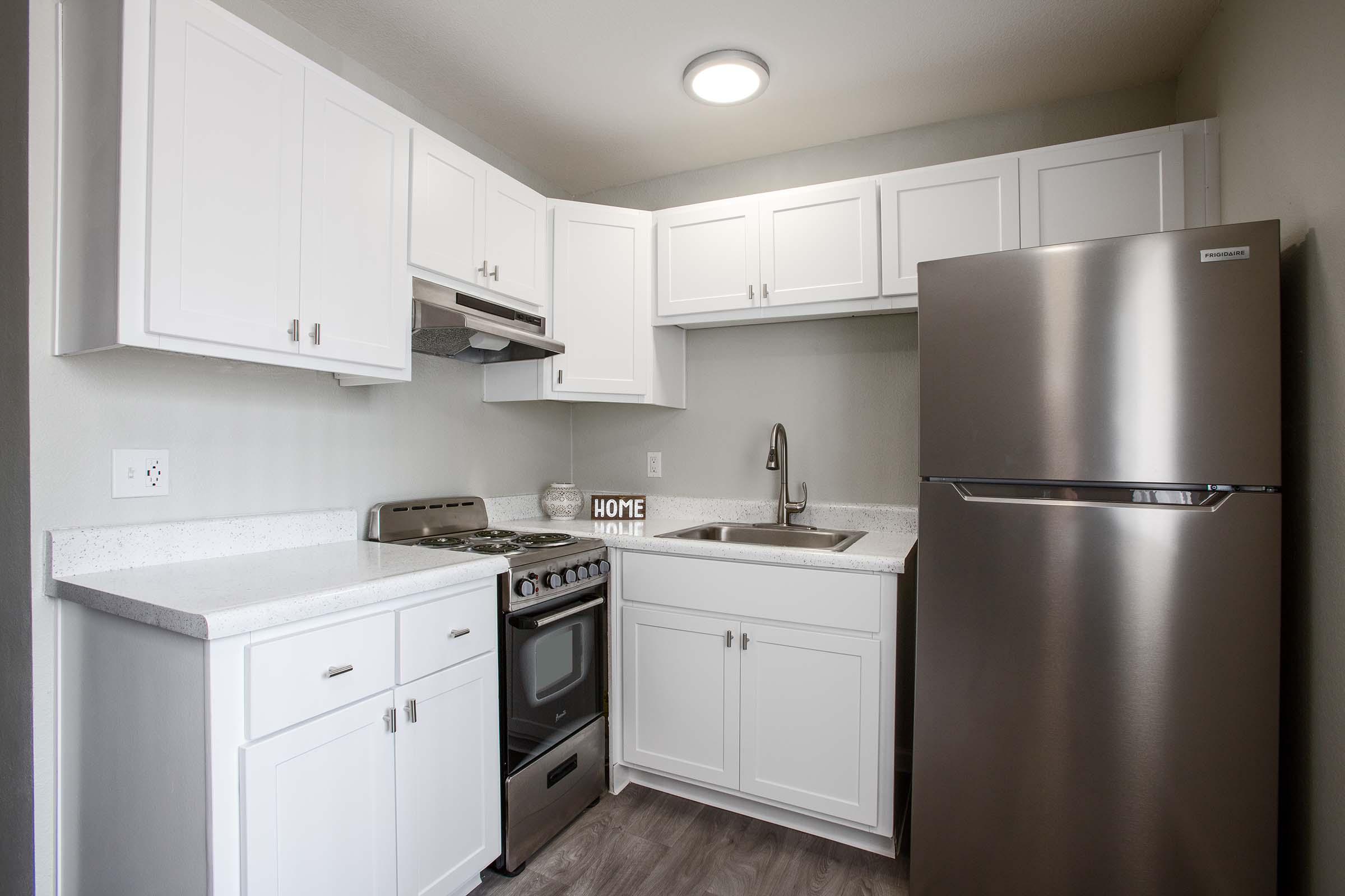 A modern kitchen featuring white cabinetry, a stainless steel refrigerator, a stove with an oven, and a sink. The countertop is light-colored, and there is a decorative "HOME" sign on the counter. The kitchen is well-lit with a ceiling light and has a clean, contemporary design.
