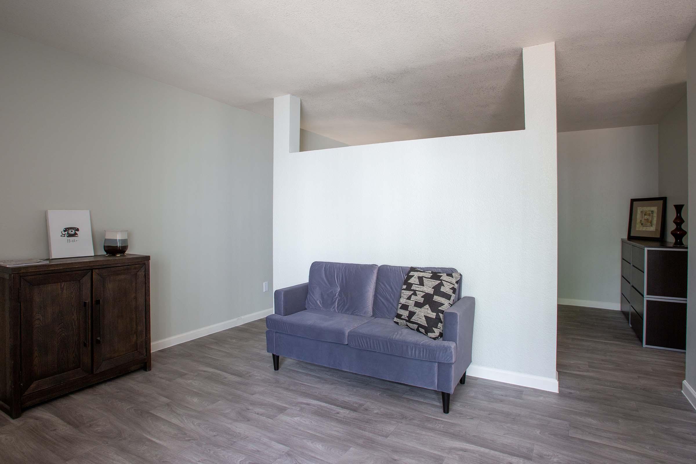 A minimalistic living room featuring a light gray wall and wooden flooring. A blue sofa with a patterned cushion is positioned centrally, with a dark wood cabinet on the left. A partition wall divides the space, and a small dresser is visible in the background. The overall ambiance is calm and spacious.