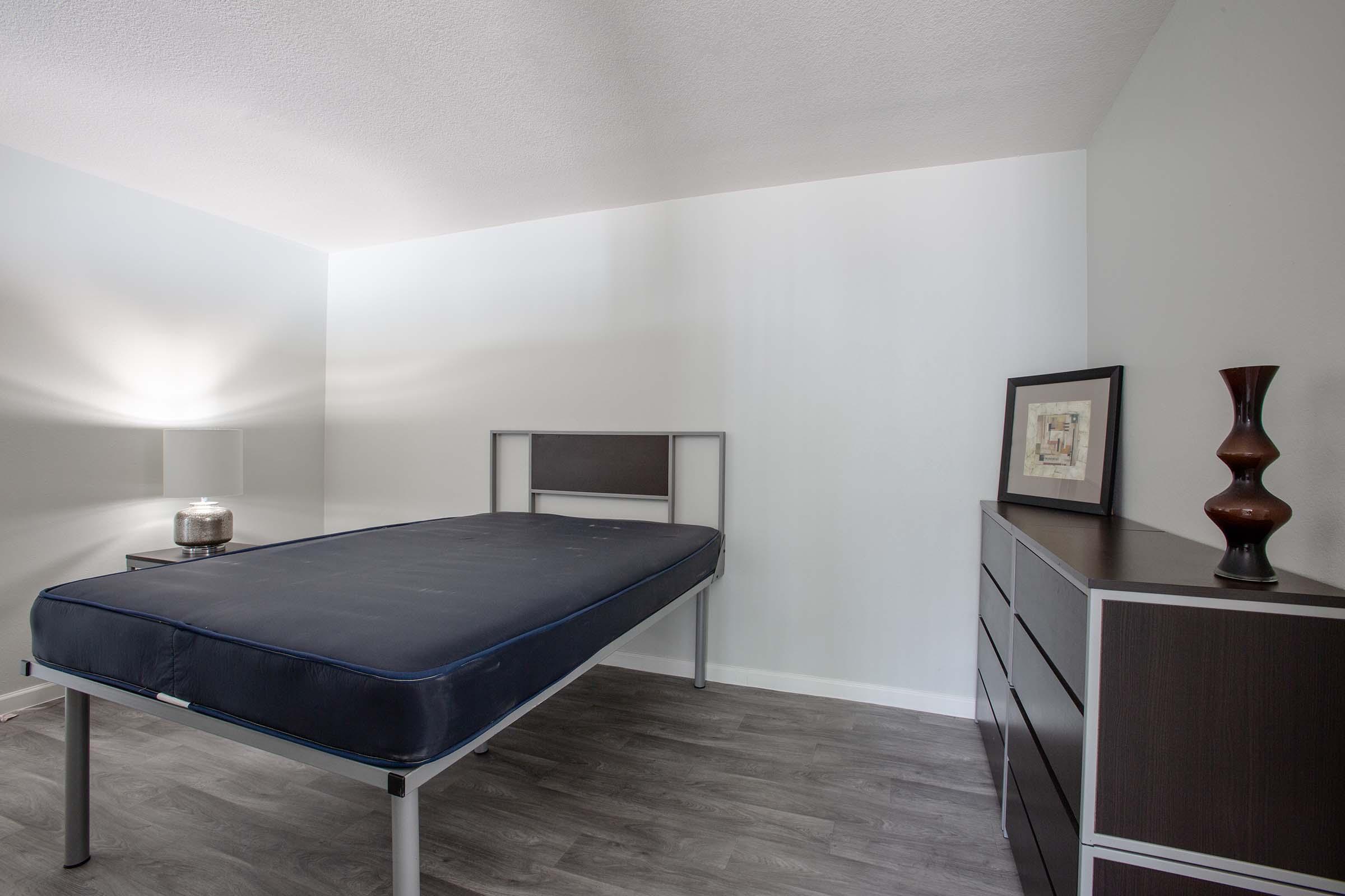A minimalist bedroom featuring a metal bed frame with a mattress, a bedside lamp, a decorative vase, and a modern dresser. The walls are painted light blue, and there is a piece of framed artwork on the wall, creating a clean and simple interior design.