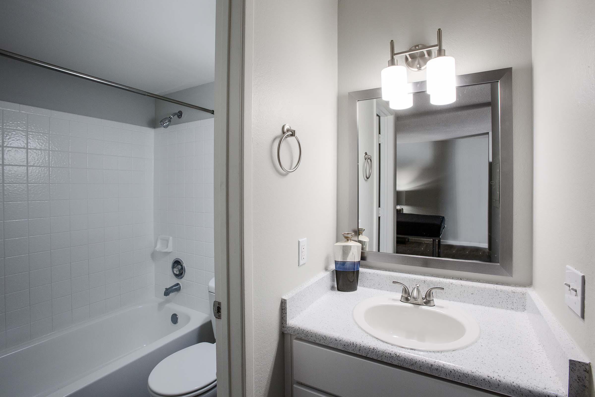 A clean and modern bathroom featuring a bathtub and shower combo, a white sink with a granite countertop, a mirrored cabinet with two light fixtures above, and a hand towel holder on the wall. The room has neutral-toned walls and a view of a nearby bathroom entrance.