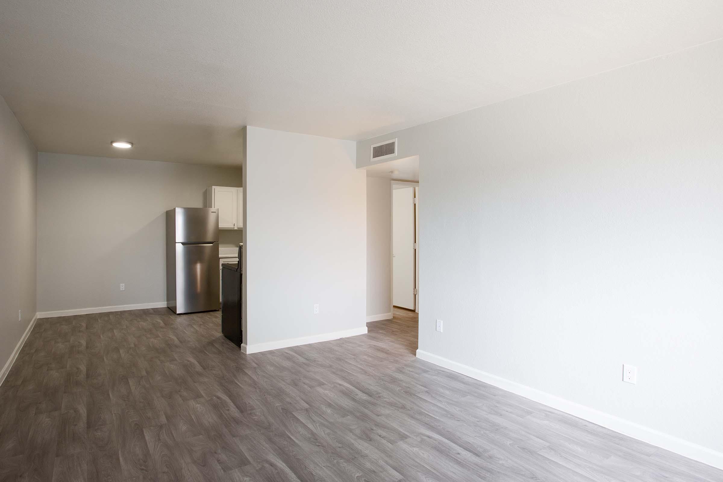 A spacious interior of an apartment featuring a modern kitchen with stainless steel appliances, including a refrigerator. The living area has light-colored walls and a wood-like floor, creating a bright and welcoming atmosphere. A doorway leads to another room, enhancing the open layout.