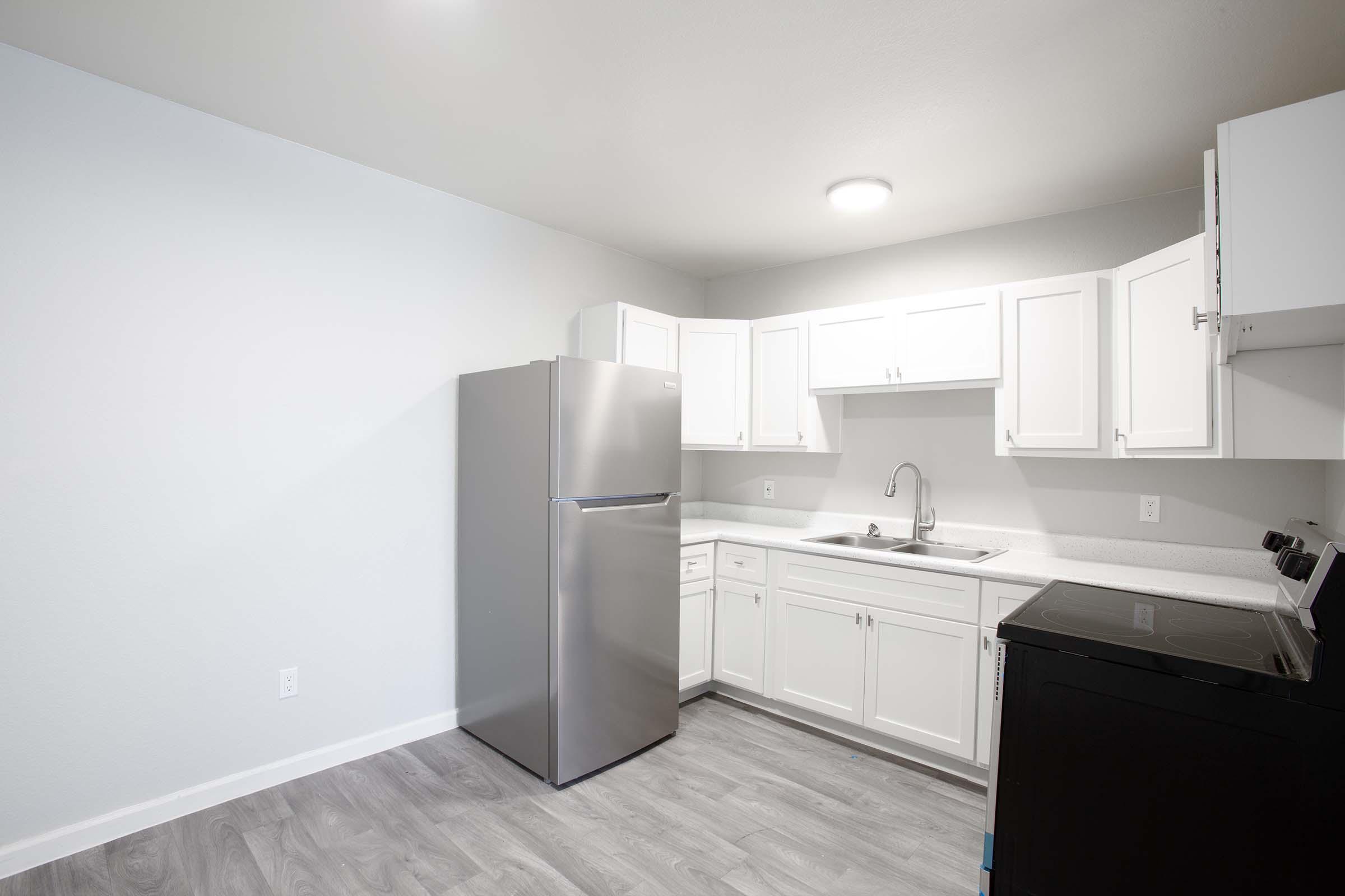 Modern kitchen featuring white cabinetry, a stainless steel refrigerator, a black stove, and a sink. The space has light-colored walls and a gray vinyl floor, creating a bright and clean atmosphere.