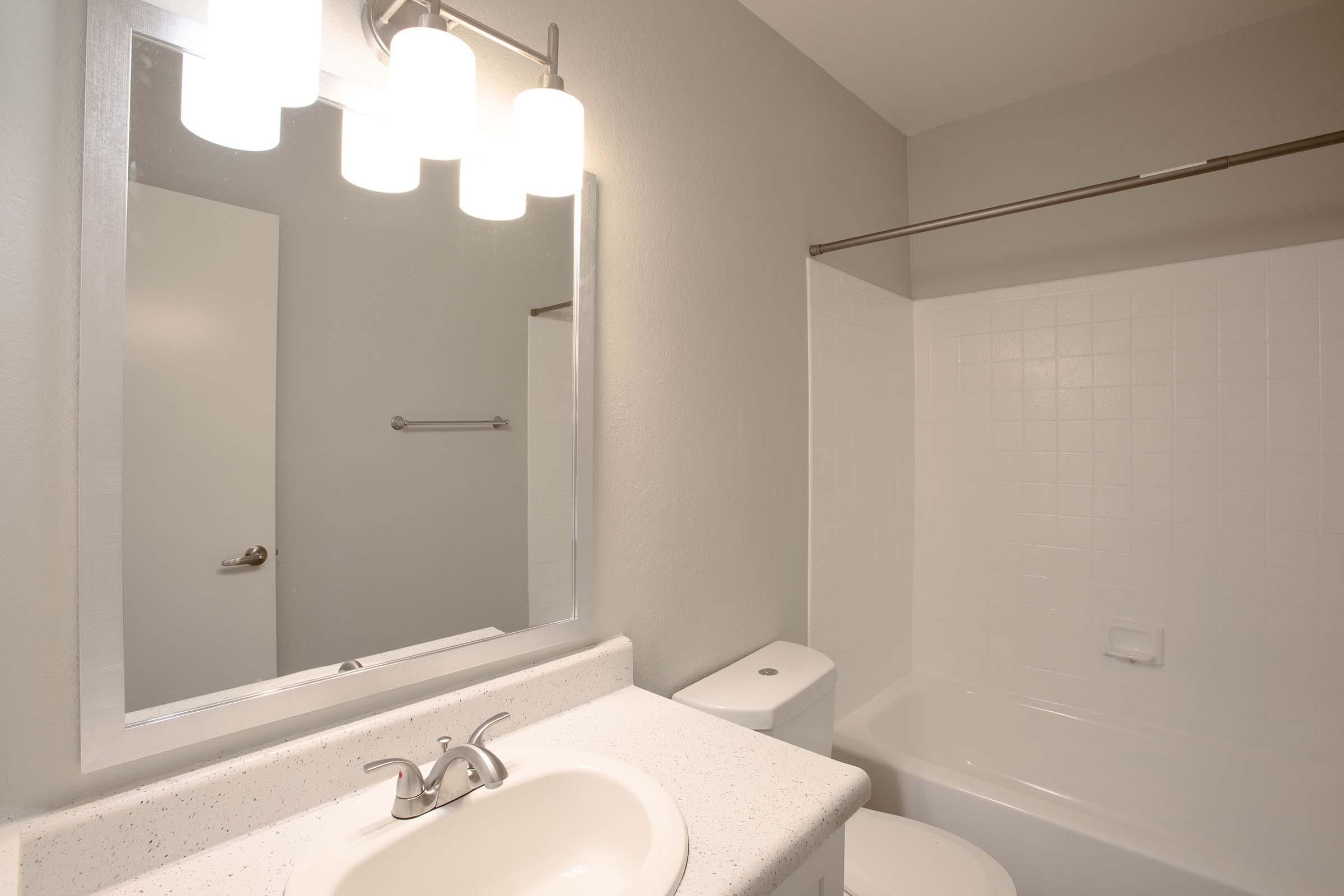 A modern bathroom featuring a white sink with a chrome faucet, a lighted mirror above the sink, and a bathtub with a shower. The walls are painted in a soft gray, and there’s a white toilet beside the bathtub. A door is visible in the background.