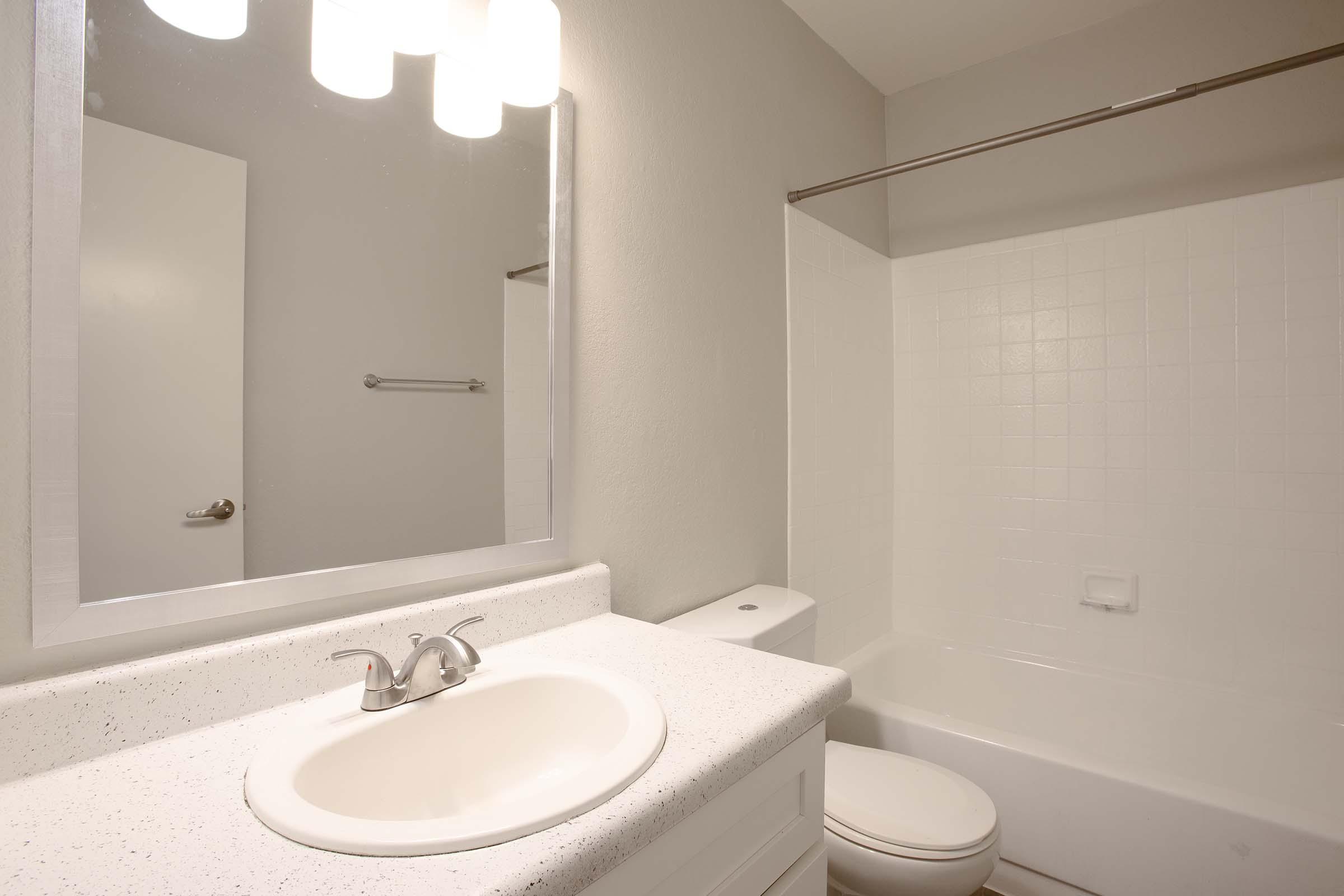 A clean and modern bathroom featuring a white sink with a shiny faucet, a large mirror above the sink, a bathtub with a shower curtain, and a simple toilet. The walls are painted in a light gray color, and there are three hanging light fixtures providing illumination.