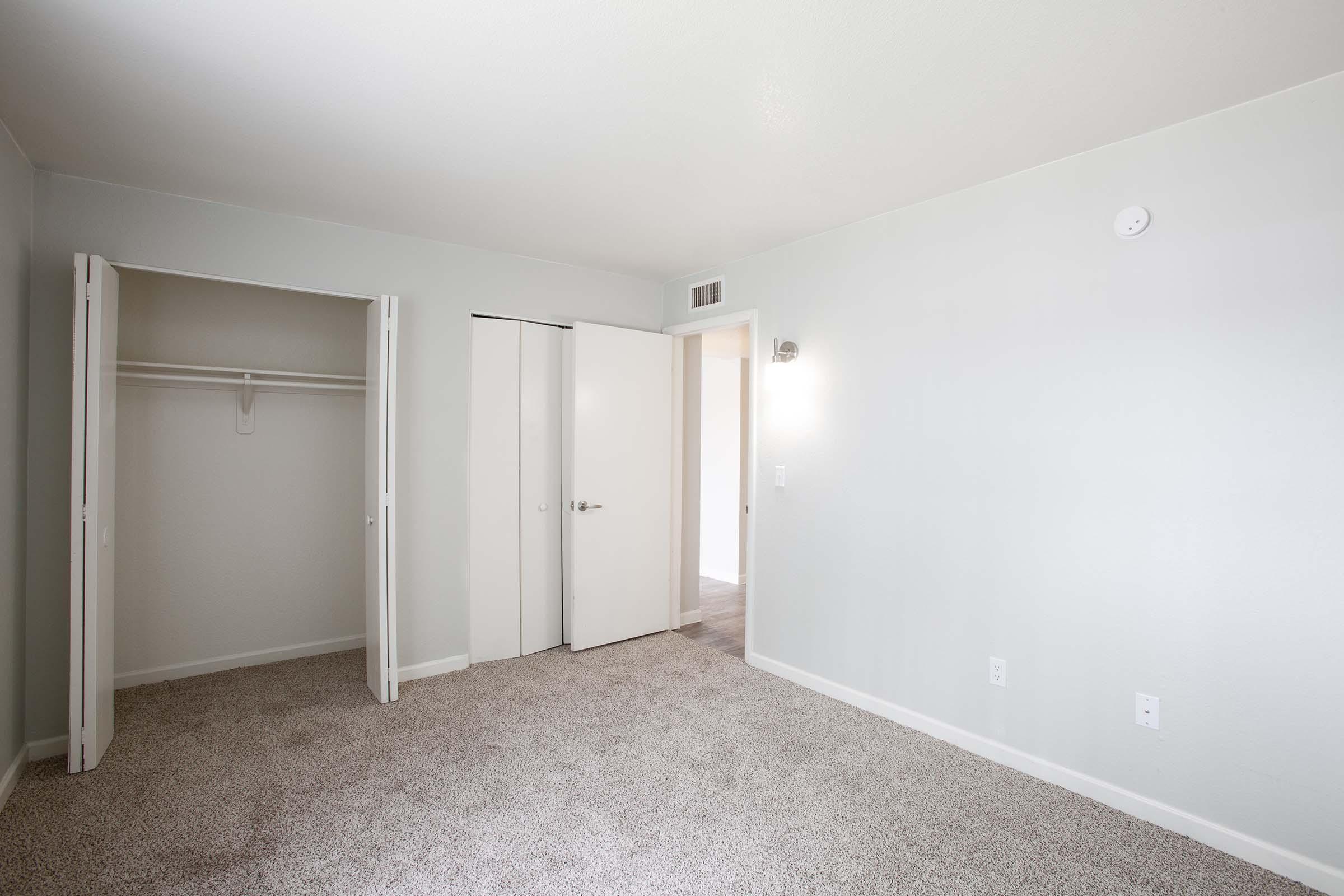 A partially furnished room with light gray walls and a carpeted floor. One side has a closet with double doors, and the opposite wall features a white door leading to another area. A wall sconce is mounted above the door, providing soft lighting. The overall ambiance is clean and minimalistic.