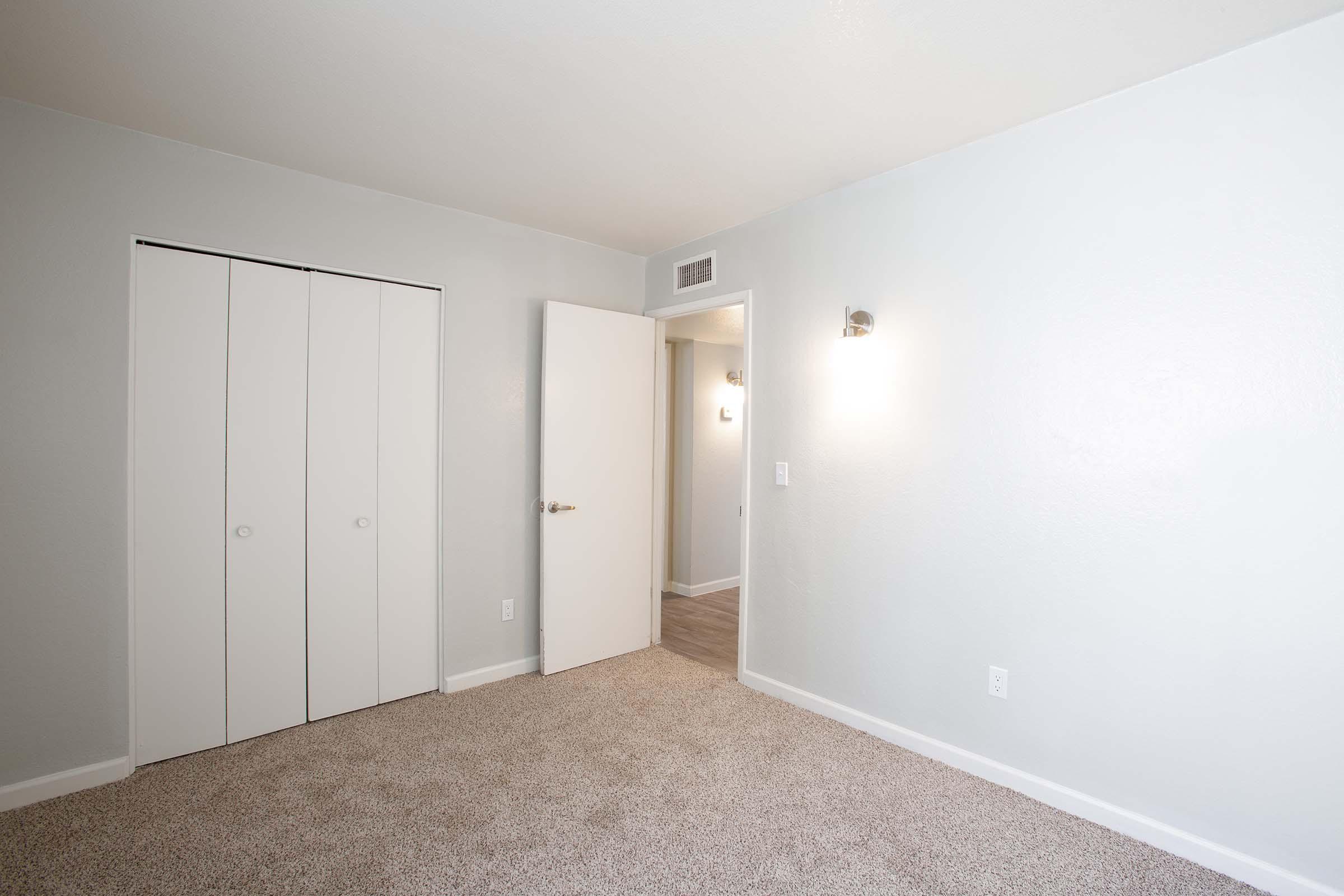 A well-lit, empty bedroom featuring light gray walls and a carpeted floor. There are two white closet doors on the left and an open door leading to another room. A wall sconce provides additional lighting. The overall feel is spacious and minimalist.