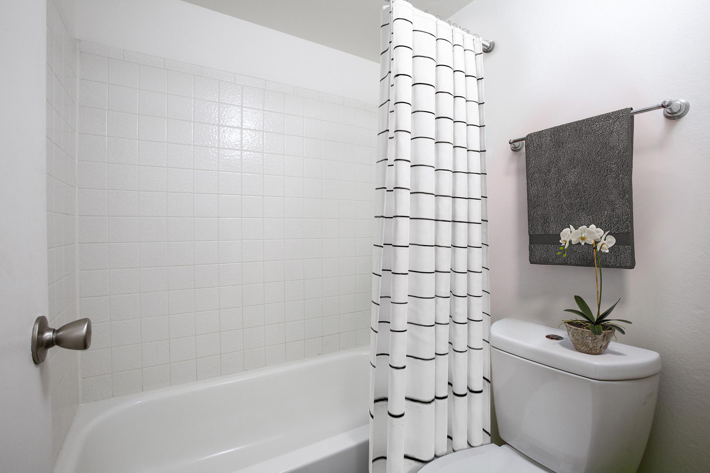 A clean, modern bathroom featuring a bathtub with a white tiled wall, a shower curtain with a patterned design, a white toilet, and a small plant with white flowers on a shelf. The decor is minimalist and fresh, creating a serene atmosphere.