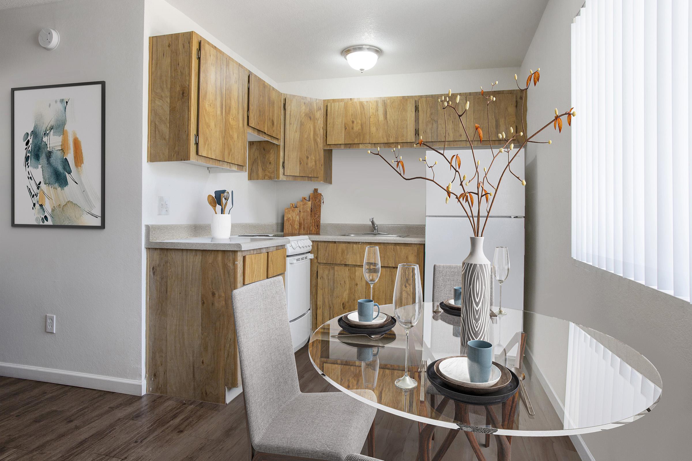 A modern kitchen with wooden cabinets and white appliances. The dining area features a glass table set with plates and glasses, accompanied by a vase of faux flowers. Bright natural light enters through vertical blinds, illuminating the minimalist decor and warm wood tones.