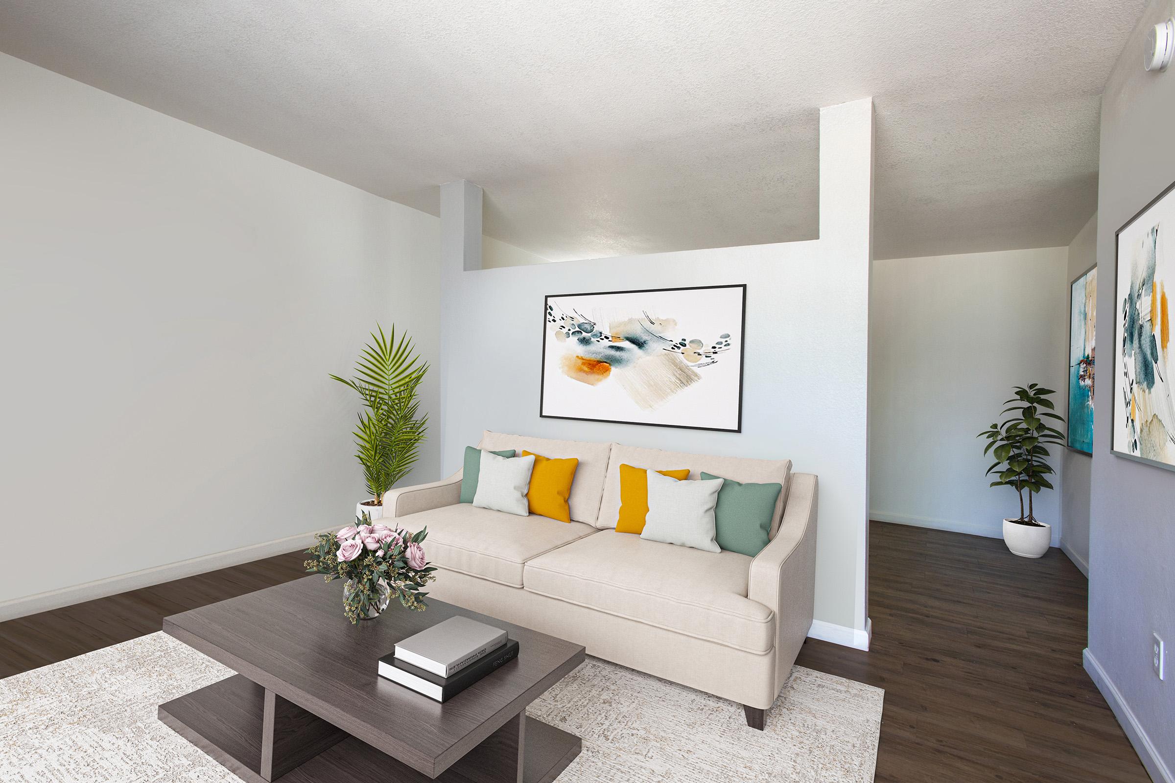 A modern living room featuring a light beige sofa with colorful accent pillows, a dark coffee table with books and decor, a lush green plant beside the sofa, and two framed artworks on the walls. The space is well-lit with neutral-colored walls and wooden flooring, creating a warm and inviting atmosphere.