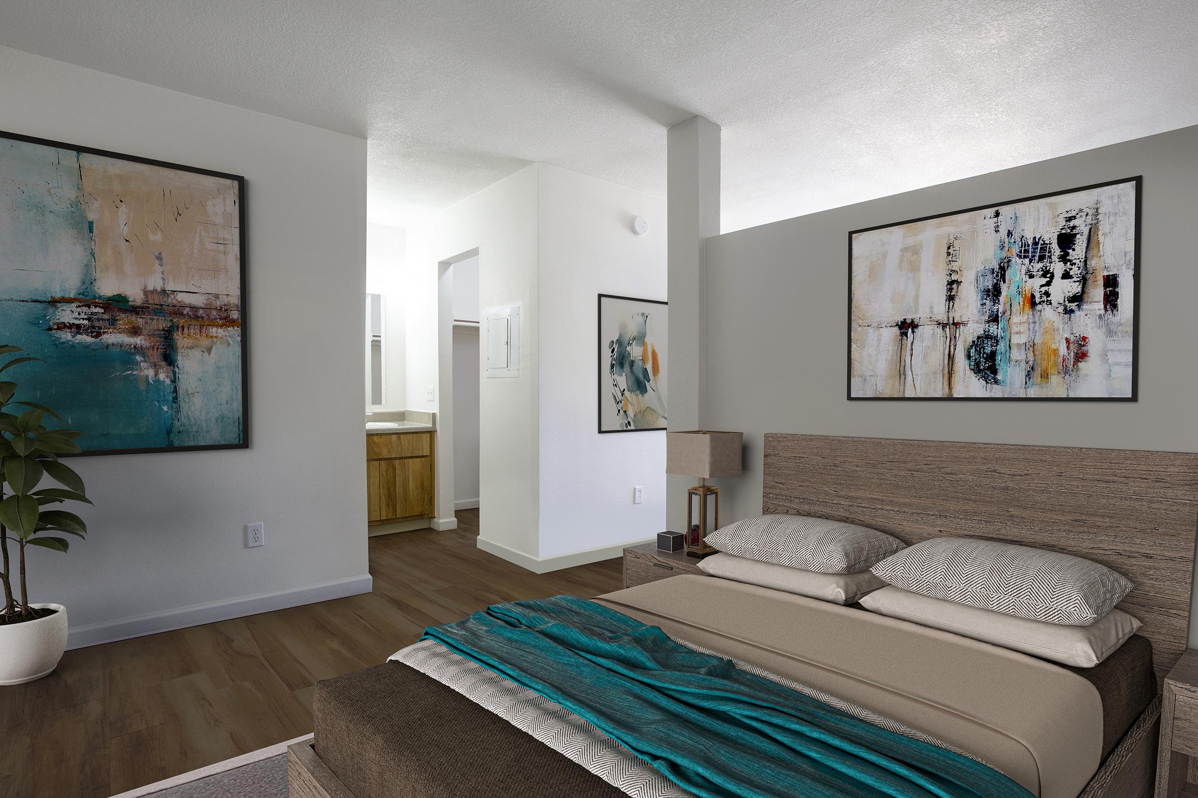 A modern bedroom featuring a large bed with neutral bedding and a teal throw. The walls are adorned with abstract paintings, and there is a potted plant beside the bed. A doorway leads to a bathroom area in the background, with light-colored cabinetry and a bright, airy atmosphere.