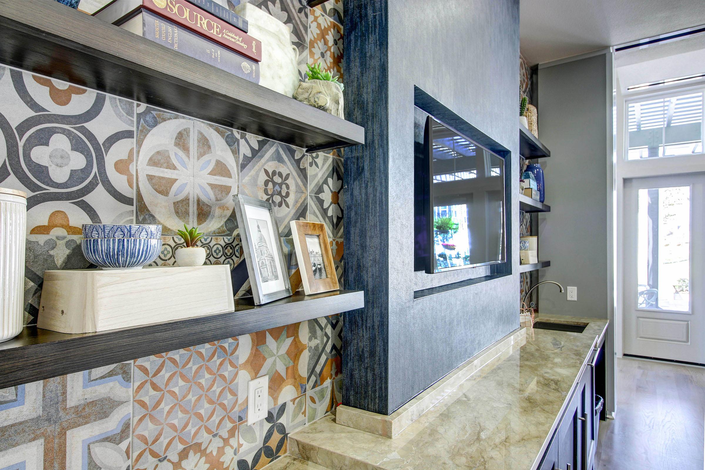 Stylish kitchen interior featuring decorative patterned tiles on the wall, dark wood floating shelves adorned with books, a small plant, and framed photos. A modern flat-screen TV is mounted above a sleek countertop, creating a contemporary and inviting atmosphere in the space. Natural light flows in from a nearby doorway.