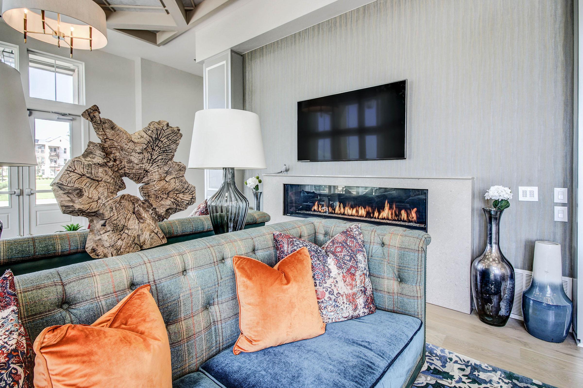A cozy living room featuring a blue and plaid sofa adorned with orange and patterned pillows. There's a modern fireplace with a sleek design below a wall-mounted TV. Decorative elements include a unique wooden sculpture, a stylish lamp, and a tall vase with flowers, creating a warm and inviting atmosphere.