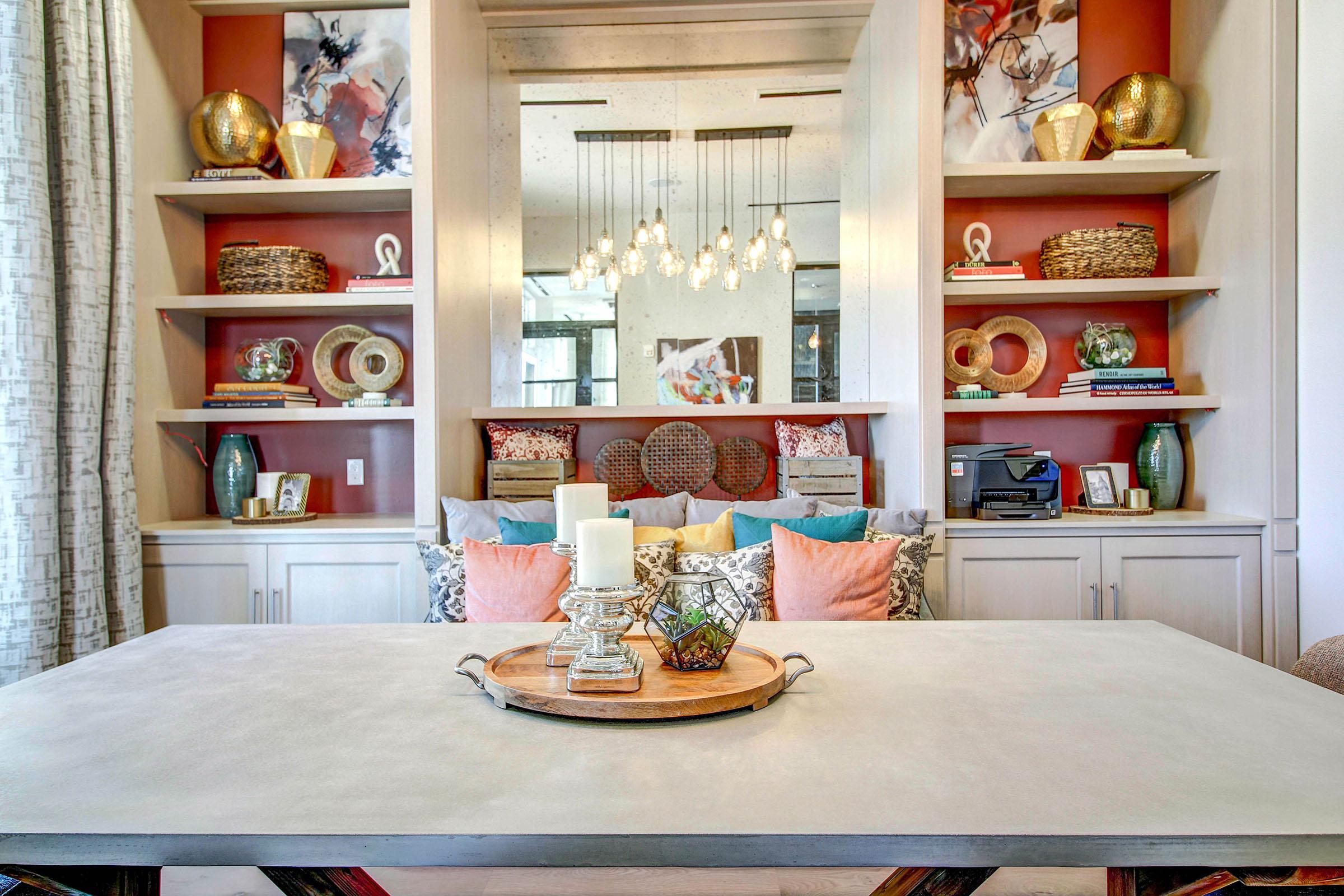 A stylish living room featuring a modern table centerpiece with a tray, candle, and decorative items. Flanking the table are shelves with decorative vases, art pieces, and woven baskets against an accented wall. Soft furnishings with colorful pillows add warmth to the inviting space.