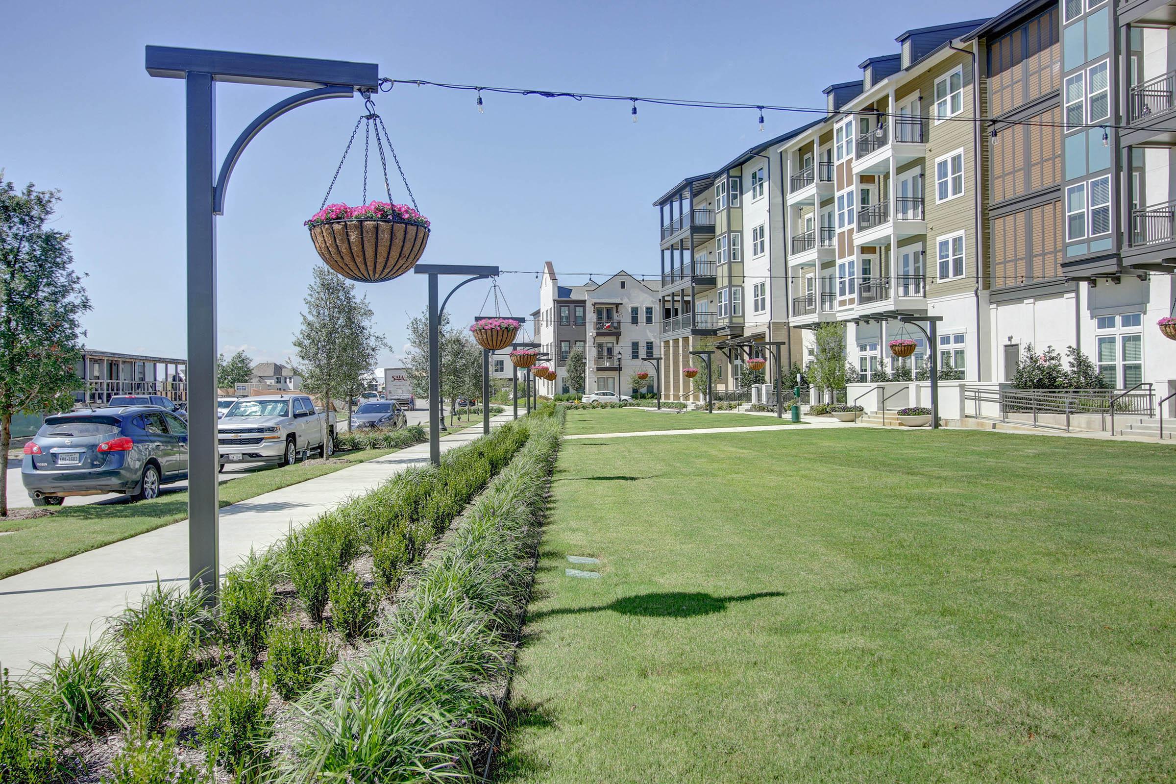 A landscaped area with a well-maintained lawn, featuring hanging flower baskets and decorative streetlights. Modern apartment buildings are visible in the background, along with parked cars along the pathway. The scene is bright and sunny, suggesting a welcoming community atmosphere.