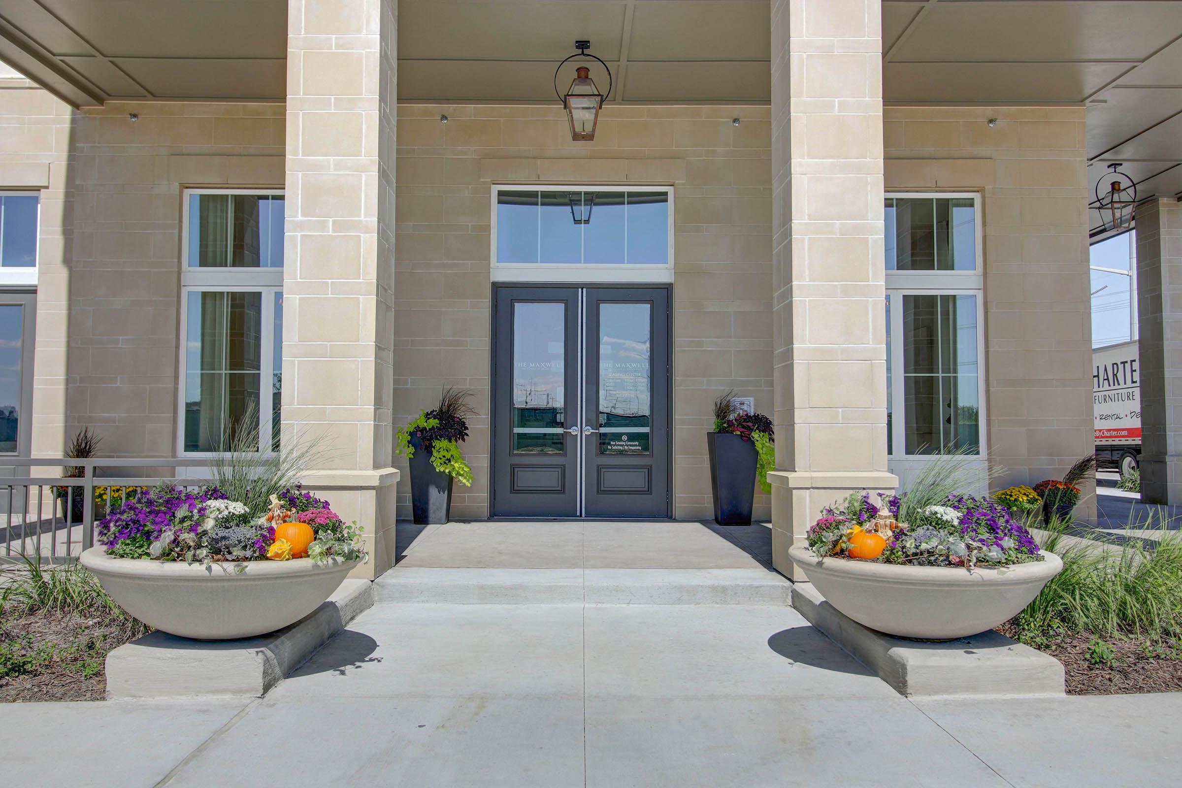 Front entrance of a building featuring large stone pillars, double doors, and planters on either side filled with colorful flowers and seasonal decorations, including pumpkins. The area is well-lit with large windows and a modern architectural design.