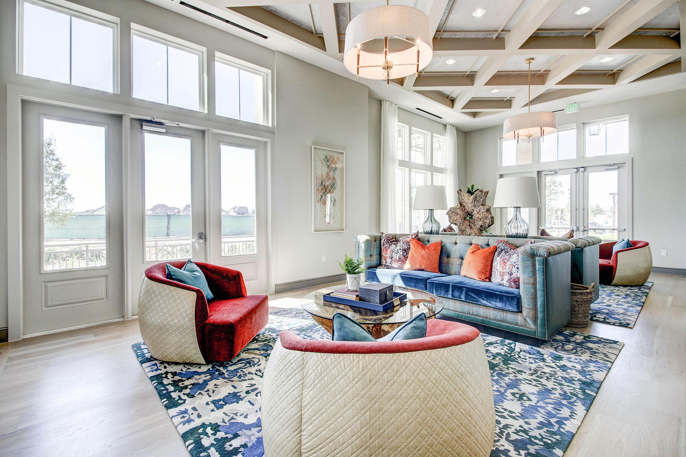 Bright and airy living room featuring large windows with natural light. The space includes a mix of colorful furniture: a blue velvet sofa, red accent chairs, and cream textured chairs. A round coffee table is centered on a patterned rug, surrounded by decorative plants and a modern chandelier.