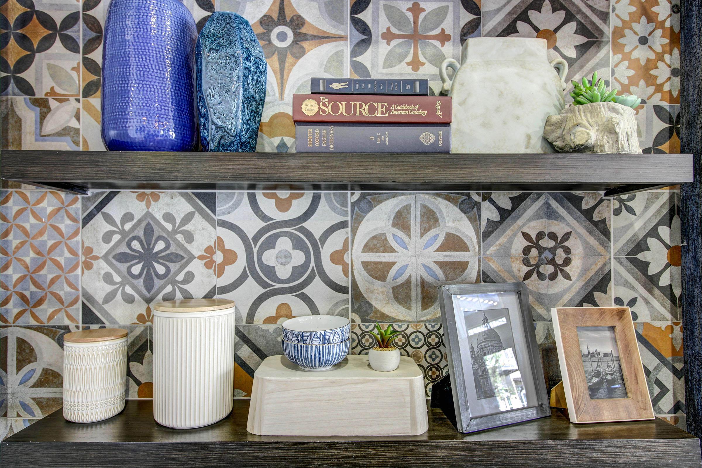 A decorative shelving unit featuring a mix of textured vases in blue, a stack of books, and various containers. There are ceramic pieces, a small planter, and picture frames, all arranged against a colorful patterned tile backdrop.