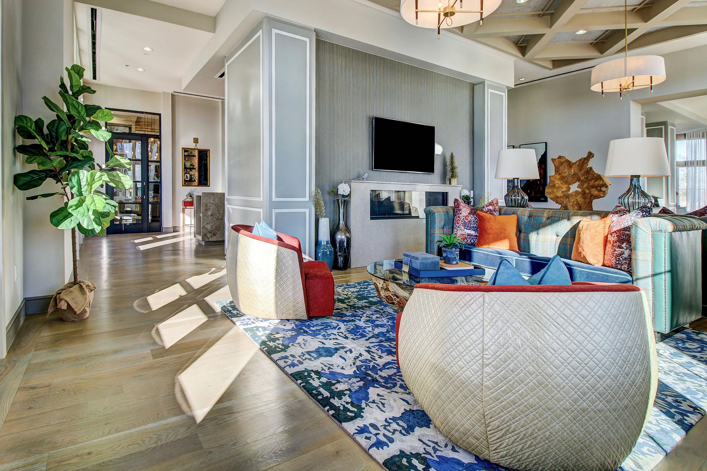 A modern living room featuring a cozy seating area with colorful pillows, a patterned rug, and a large TV mounted on the wall. Bright, natural light filters through large windows, highlighting plants and stylish decor elements in a spacious, inviting layout.